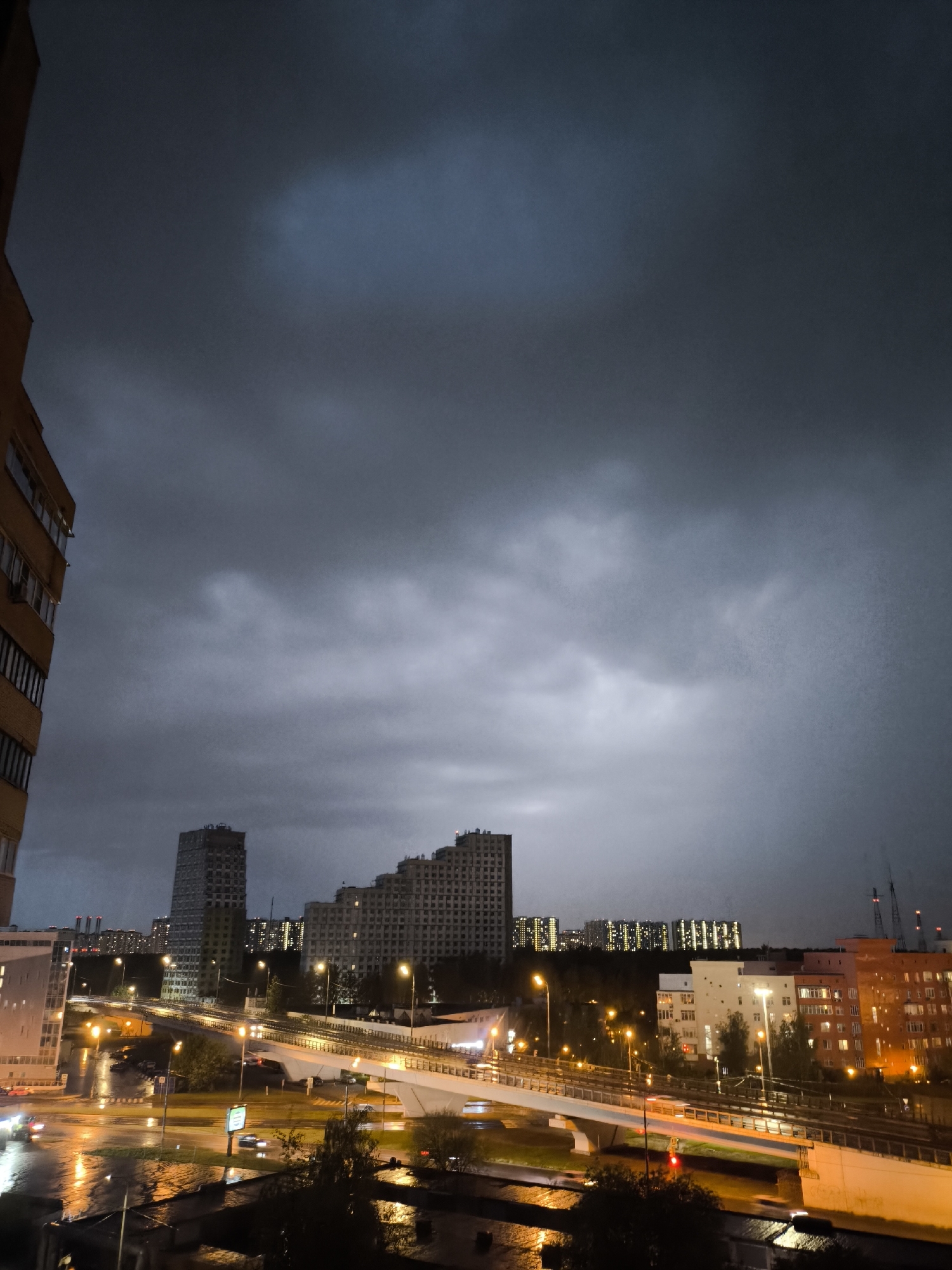 The image depicts a nighttime cityscape under a cloudy sky. In the foreground, a wet road reflects the streetlights and car headlights, creating bright, blurred lines. A bridge structure crosses the road, illuminated with yellow lights. Beyond the bridge are rows of apartment buildings, some of which appear to be high-rise, with lit windows. The sky is predominantly dark with varying shades of grey, indicating heavy cloud cover. A portion of a building is visible on the left side of the frame. Cranes are visible in the distance, suggesting ongoing construction. The location appears to be an urban area at night during or after rainfall.