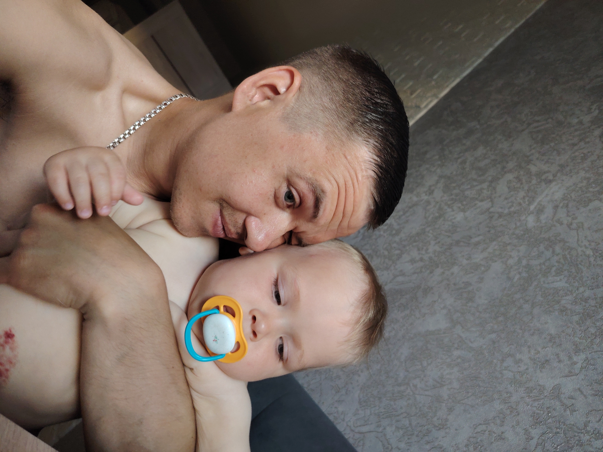 A shirtless man with short, dark hair and a silver chain necklace is holding a baby with a pacifier in its mouth. The man is looking down at the baby with affection. The background is a gray wall. The location is inferred to be in the city of Chaikovsky, Russia.