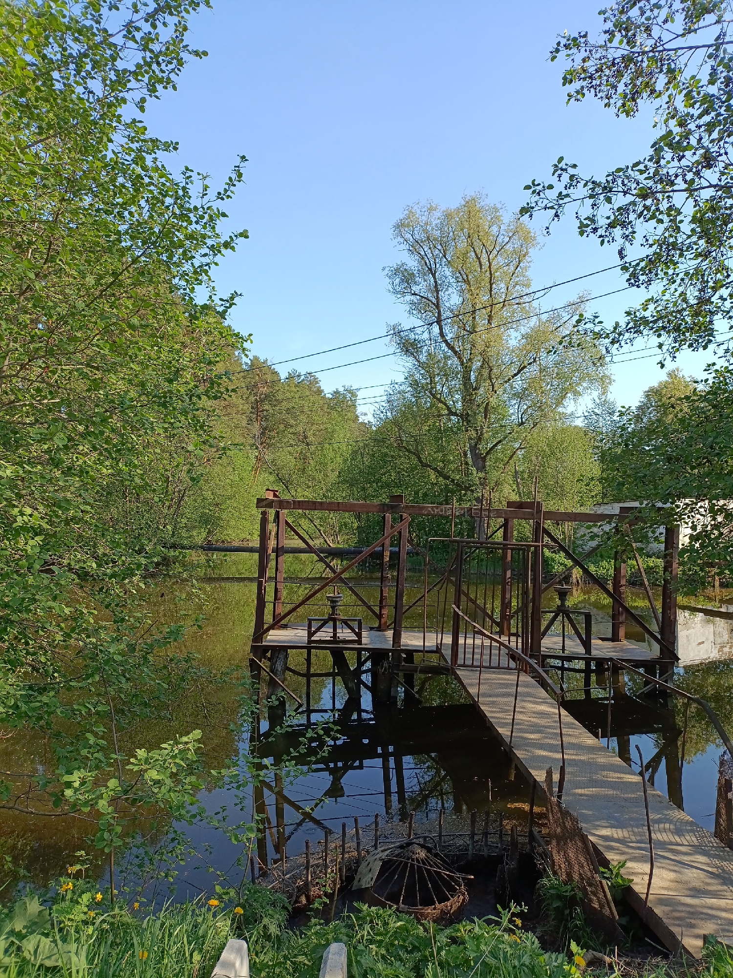 The image depicts a small dam or water control structure situated on a pond or small lake, surrounded by trees.
The dam consists of a metal framework with wooden platforms. A metal bridge leads to the structure, suggesting a means of access for maintenance or operation.
The water is dark and still, reflecting the surrounding trees and the structure. The vegetation appears lush and green, indicating a well-watered environment.
The sky is clear and blue. The photo was taken during daylight.
Although the location is said to be Chaikovsky, Russia, there are no prominent city-specific indicators to clearly determine the location.