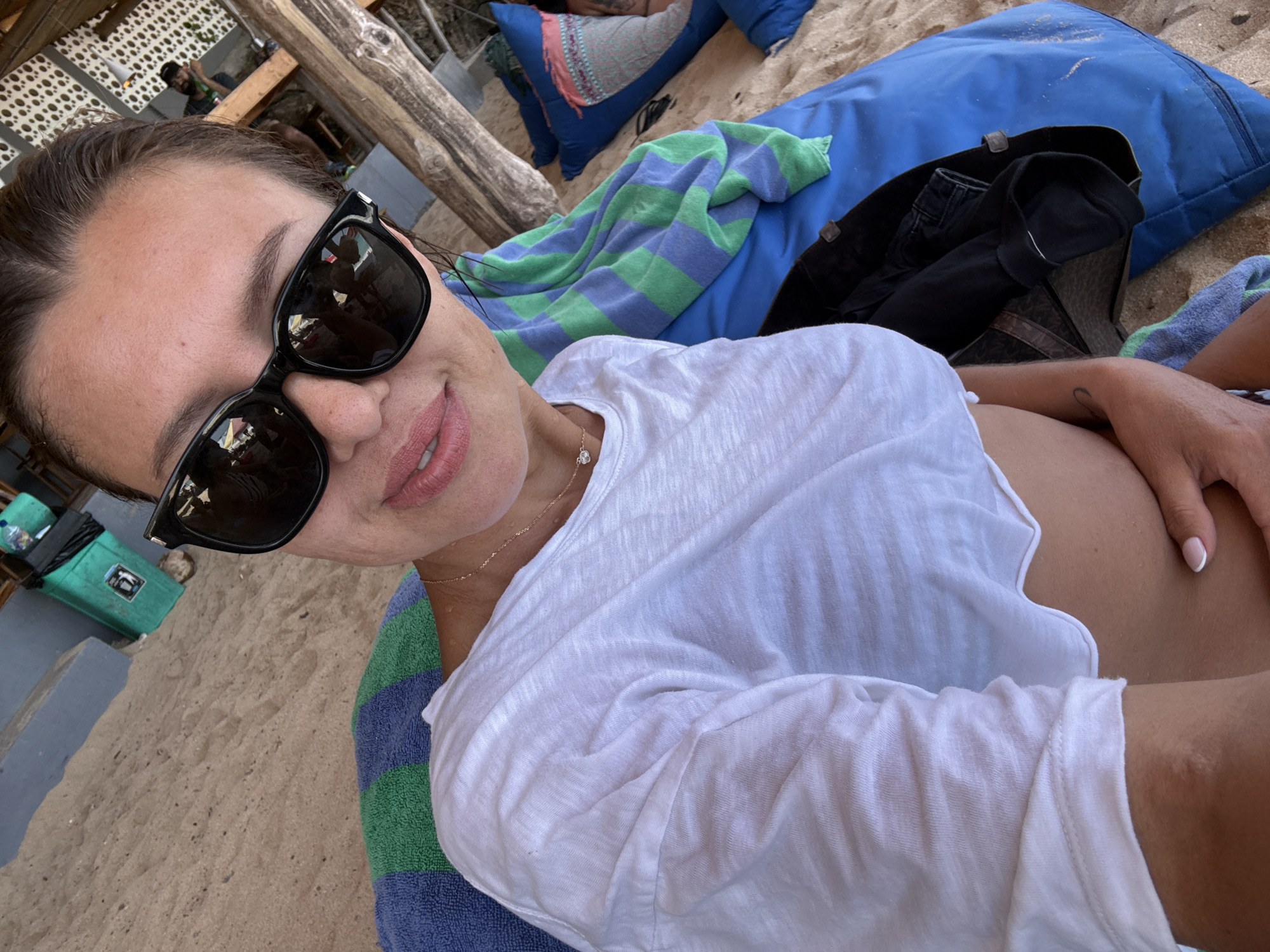 The image is a selfie taken on a beach in Kuta Selatan, Indonesia. A woman wearing sunglasses and a white shirt is lying down, holding the camera. The background shows a sandy beach, beach furniture (blue cushions, a blue and green striped towel), and wooden structures. A trash can can be seen and a man is visible further in the background under a shaded structure.