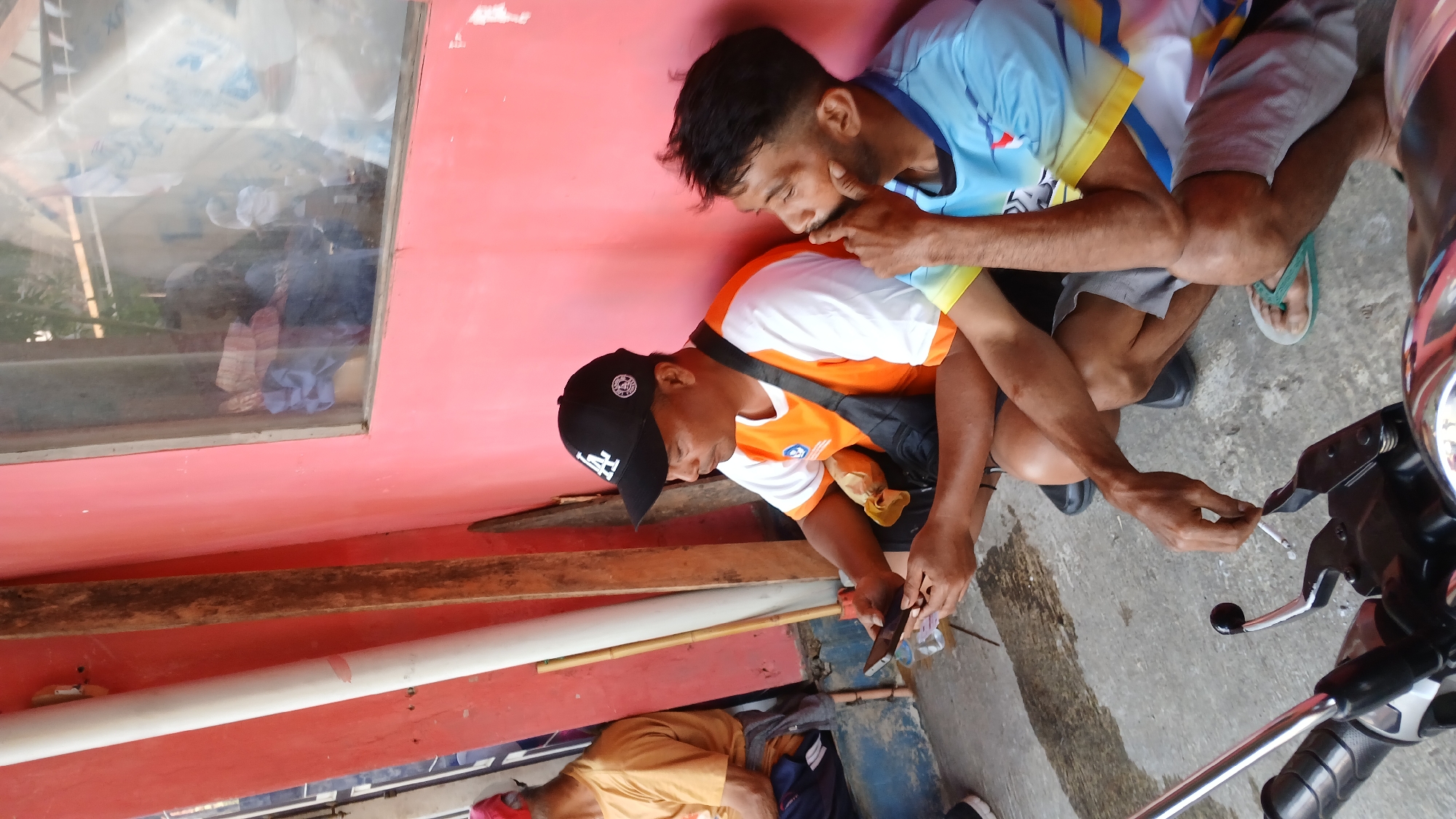 Two men are crouching on a concrete floor next to a pink wall. The man on the left is wearing a black baseball cap with a white "LA" logo, an orange and white t-shirt, and a black crossbody bag. He is holding a smartphone in his hands, looking down at it. Beside him on the ground is a piece of yellow fabric, possibly food. The man on the right is wearing a blue and yellow t-shirt and grey shorts. He is resting his chin on his hand, looking to his right. In front of the two men, a motorcycle is partially visible. To the left of the men, a white pipe and a wooden pole are leaning against the pink wall. The environment appears to be an outdoor, possibly covered, area, suggesting a casual gathering place. The image was taken in the city of Mendut, Indonesia.