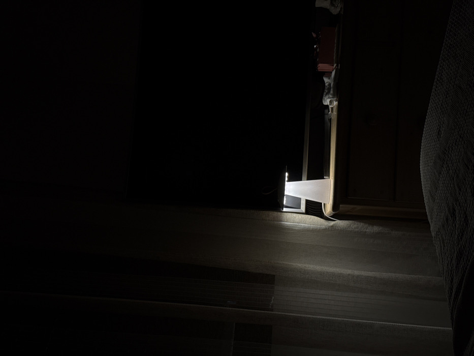 The image displays an interior room, likely a bedroom, located in Dos Hermanas, Spain, under low-light conditions. The foreground features a dark, textured blanket partially covering a bed. To the left, a light-colored, sheer curtain hangs, obscuring a window or wall. In the midground, a wooden dresser with visible drawers supports a white, conical-shaped lamp with a flat top, which is illuminated and emitting light downwards and forwards. A white power cord extends from the lamp's base. Behind the lamp, a large, dark, rectangular object, consistent with a television or monitor screen, is visible. To the right of the lamp on the dresser, a red box-like object and several light-colored fabric items are present. The background and upper portions of the room are obscured by darkness. No individuals are visible.