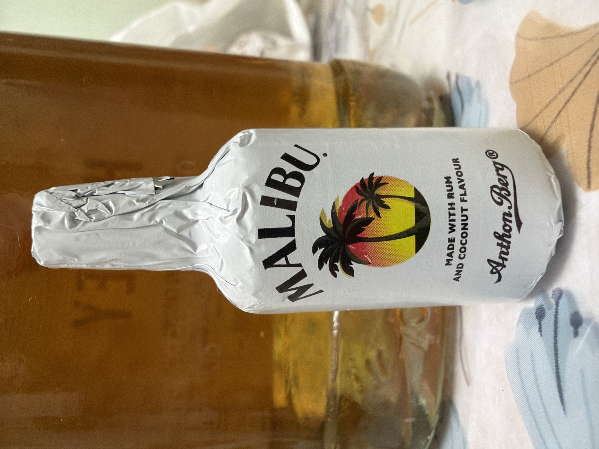 A small, foil-wrapped bottle of Malibu rum with coconut flavor is the primary subject of the image. The bottle is standing upright on a patterned surface, possibly a tablecloth. The foil is silver and has a textured appearance. The label on the bottle features the "Malibu" logo in black, followed by an illustration of palm trees against a sunset, with the text "MADE WITH RUM AND COCONUT FLAVOUR" and "Anthon Berg" below. In the background, a larger glass container filled with a golden-brown liquid is visible, its contents appearing somewhat cloudy. The lighting in the image is soft, creating a slightly diffused atmosphere. The scene is indoors, suggested by the presence of objects and a patterned surface. The image does not provide any information about the city of Kyiv, Ukraine, or any specific location within it. There are no people or actions depicted in the image.