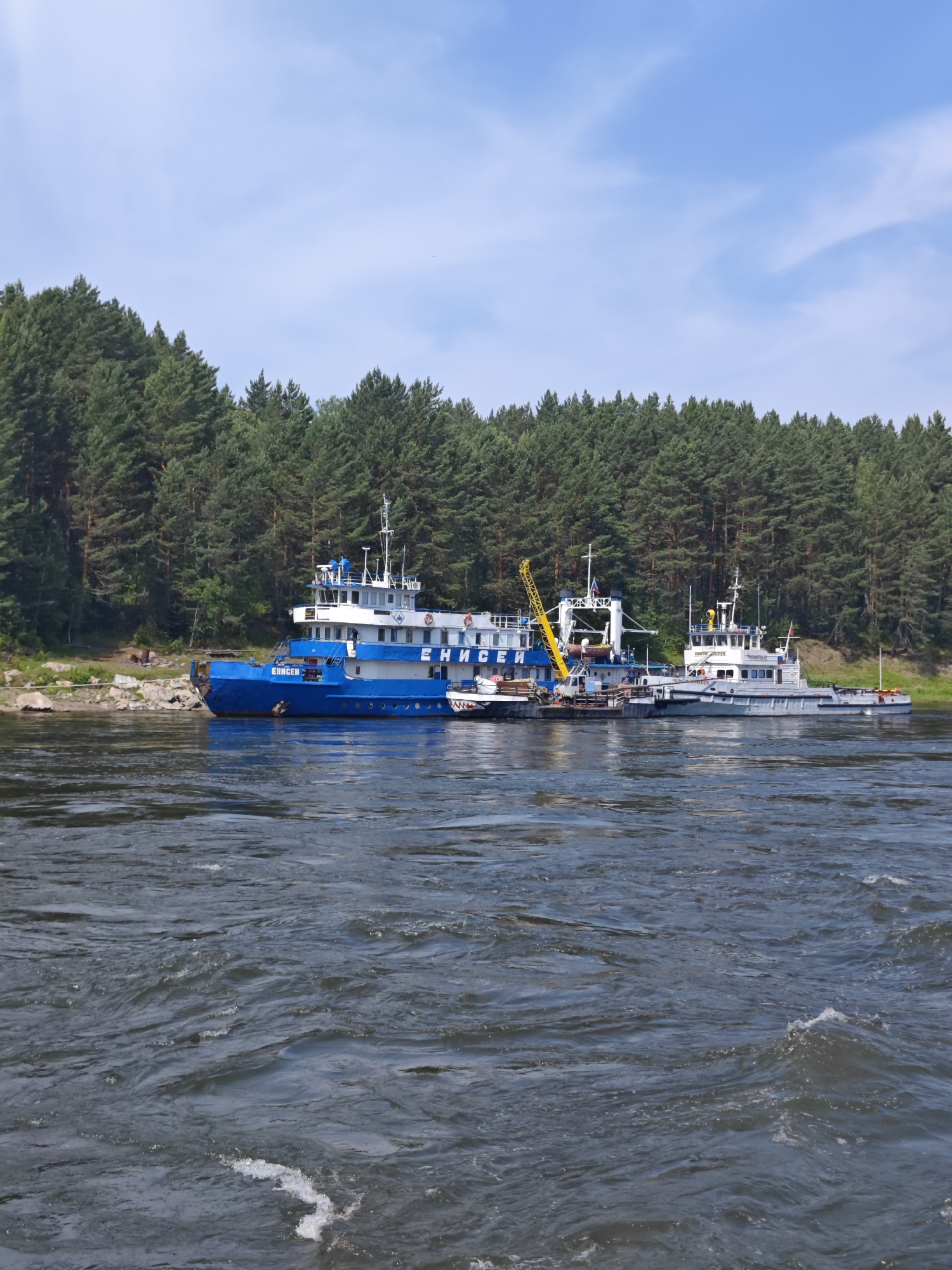 A blue and white vessel, identified by the inscription "ЕНИСЕЙ" (Yenisei) on its side, is docked on the right bank of a wide, turbulent river. Adjacent to it is a smaller, gray vessel. Both are situated in front of a dense, dark green coniferous forest that stretches across the horizon. The sky above is a clear blue with scattered white clouds. The river water is dark and shows signs of strong currents and ripples. The scene appears to be in the city of Porog, Russia, as suggested by the context.