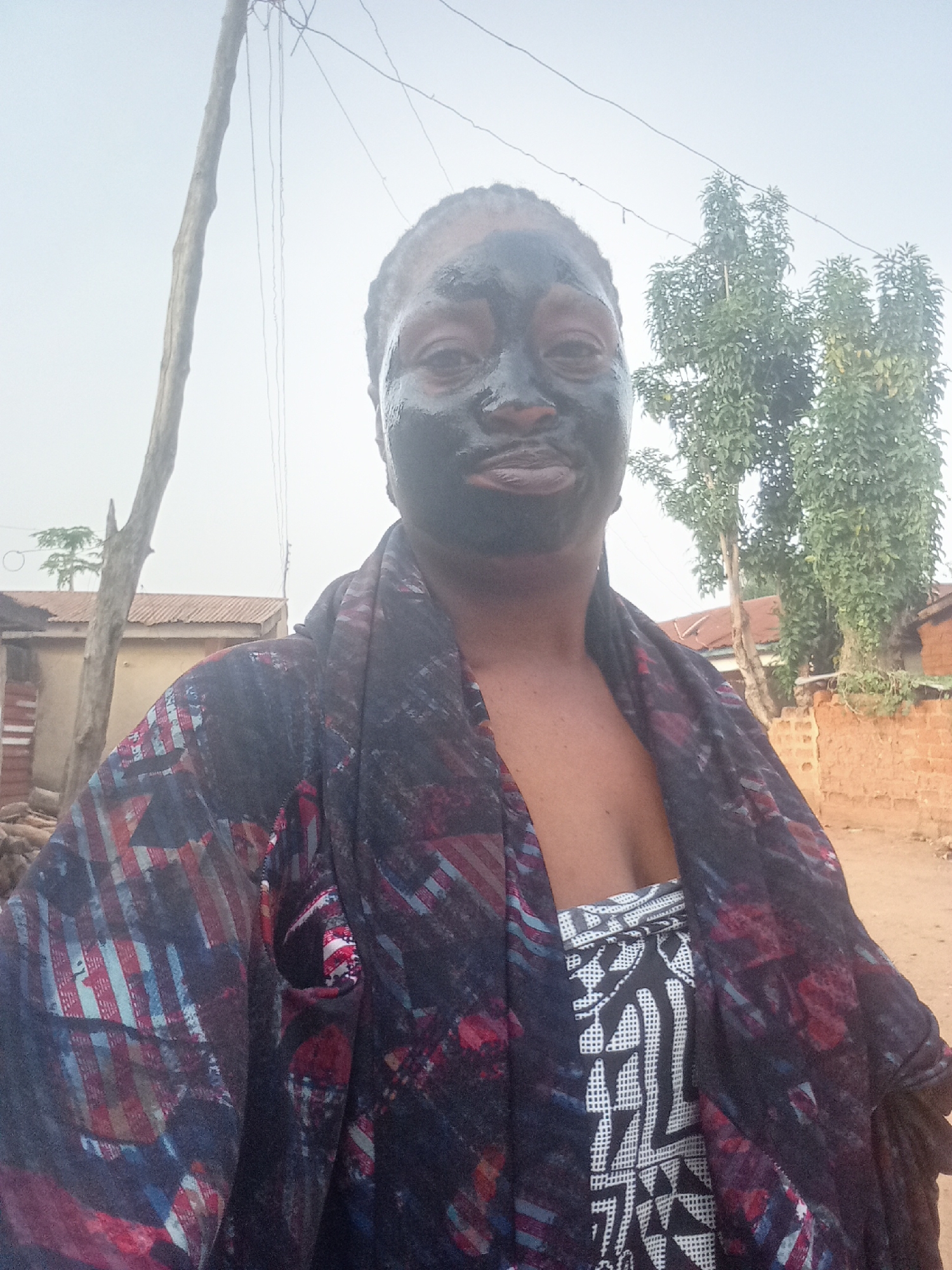 A medium shot captures a person with a dark facial mask. The individual is wearing a dress with a geometric black and white pattern and a multi-colored scarf. The background features a rural or semi-urban landscape. Elements of the environment include modest buildings with rusted roofs, a brick wall, and a tree with dense foliage. A wooden pole with wires is also visible. The image suggests an outdoor setting in Akwanga, Nigeria, or a similar location.