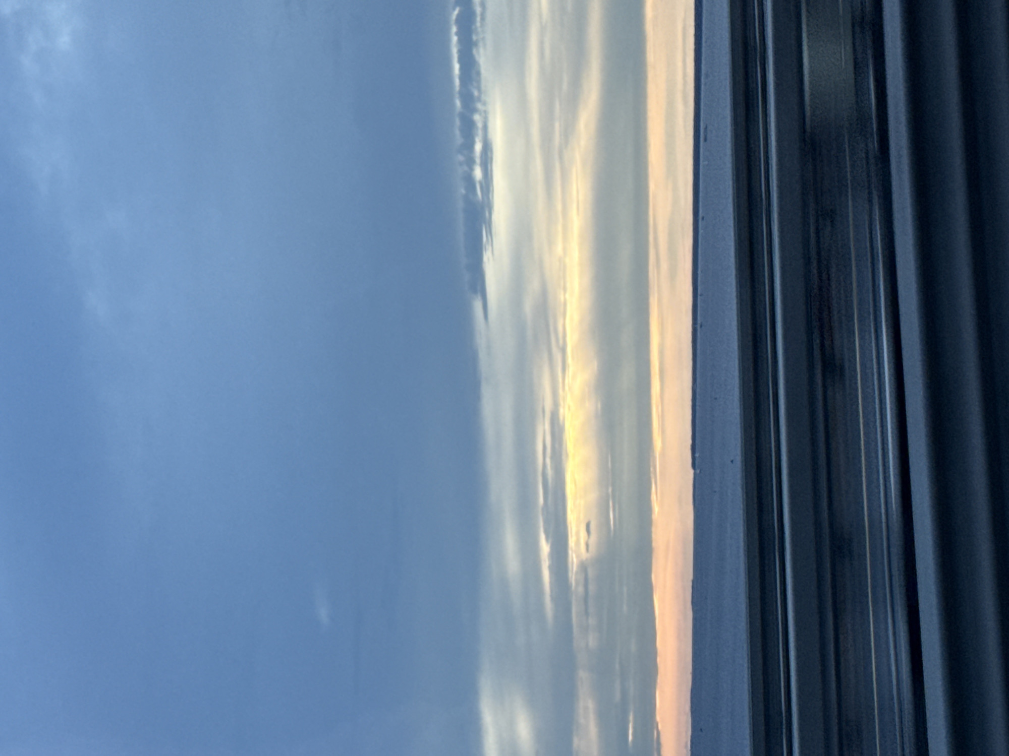 The image depicts a wide, calm body of water stretching to the horizon under a cloudy sky. The sky is a mix of deep blue and lighter gray clouds, with streaks of yellow and orange light breaking through in the middle distance, suggesting either sunrise or sunset. A faint, hazy landmass is visible on the horizon. In the foreground, several horizontal metal barriers or railings are visible, creating a layered effect that suggests the photo was taken from a bridge or elevated roadway. The overall impression is one of a serene, expansive natural landscape, viewed from an urban or semi-urban vantage point. The presence of the barriers strongly implies the location is on a bridge over a body of water in Saint Petersburg, Russia, given its numerous bridges and waterways. However, without specific landmarks, pinpointing the exact location within the city is not possible from this image alone. There are no people or active events depicted in the scene.