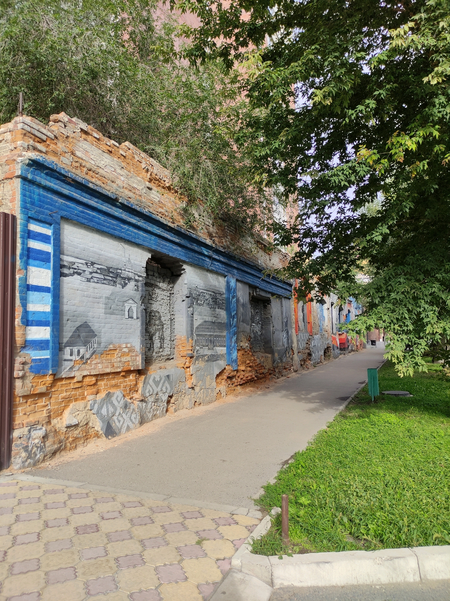 The image depicts an outdoor scene with a dilapidated brick wall on the left, adorned with a mural. The mural features architectural drawings in grayscale with blue accents. The wall is partially crumbling, revealing older bricks and weathered plaster. To the right of the wall is a paved walkway, bordered by a strip of lush green grass. Further to the right, large trees with dense foliage overhang the walkway and grass, casting shadows. In the background, there's a glimpse of more buildings and greenery. A green trash receptacle is visible on the grass. The foreground shows a patterned paved sidewalk. This scene is located in the city of Semey, Kazakhstan.