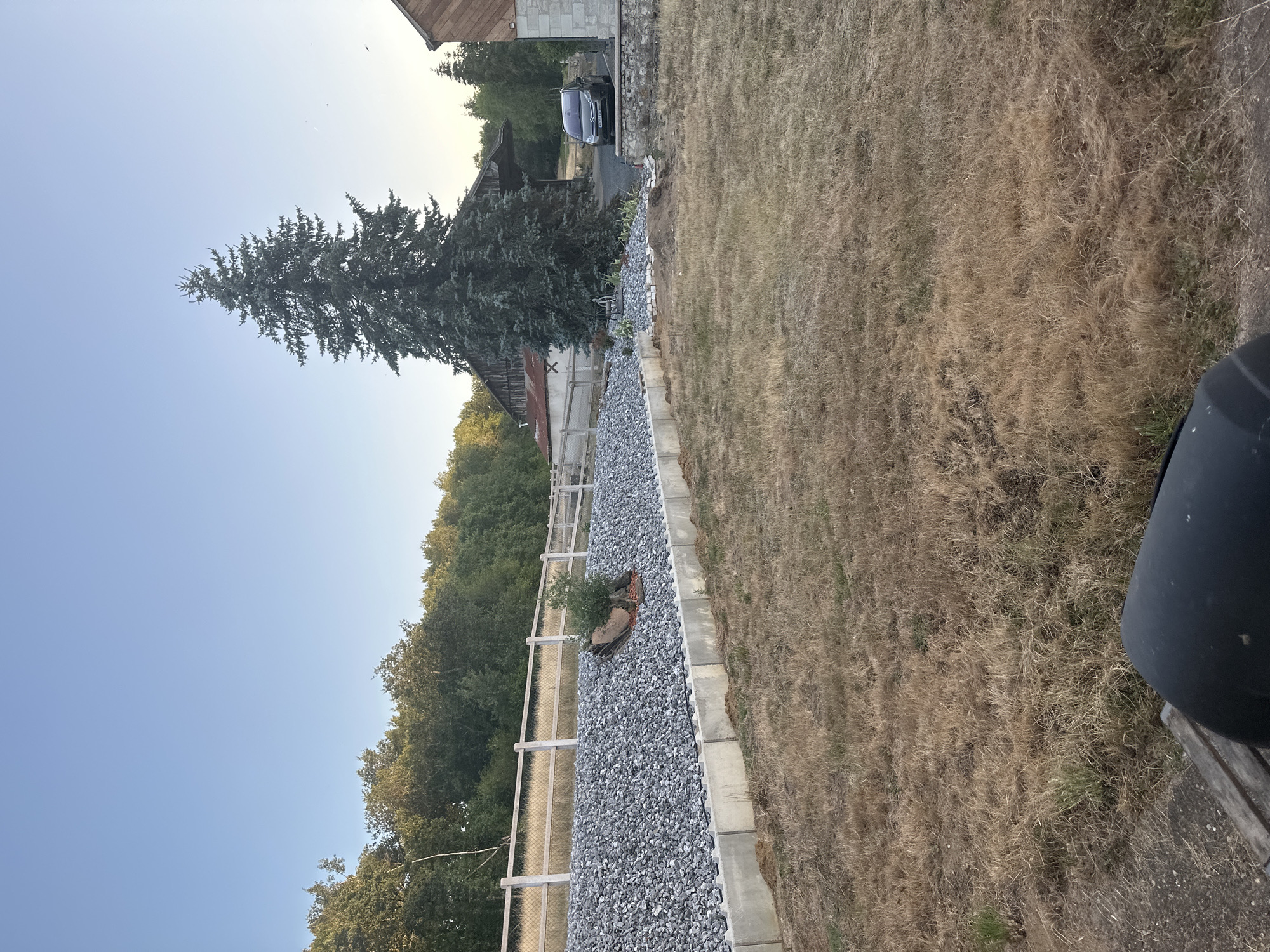 The image depicts an outdoor scene, likely in Bourgueil, France, featuring a rural landscape with elements of construction and a natural environment. A large, mature evergreen tree stands prominently in the center-right of the frame, positioned near a rustic wooden barn with a corrugated metal roof. To the right of the barn, a section of a stone building is visible. In the foreground and midground, dry, yellowed grass covers the ground, suggesting a dry season.
A retaining wall, constructed from concrete blocks, runs along the left side of the image, supporting a slope covered with light gray gravel. A white wooden fence with concrete posts runs along the top of the gravel slope, demarcating the property line or a pathway. A small, green bush is planted on a rocky outcrop on the gravel slope.
A dark-colored car, a Citroën C4 Picasso, is parked on a gravel driveway in front of the barn. The sky is a clear, pale blue, indicating fair weather. The overall impression is one of a recently developed or altered landscape in a rural setting, possibly a farm or residential property, with a blend of natural elements and human construction. There are no people or animals visible in the image.