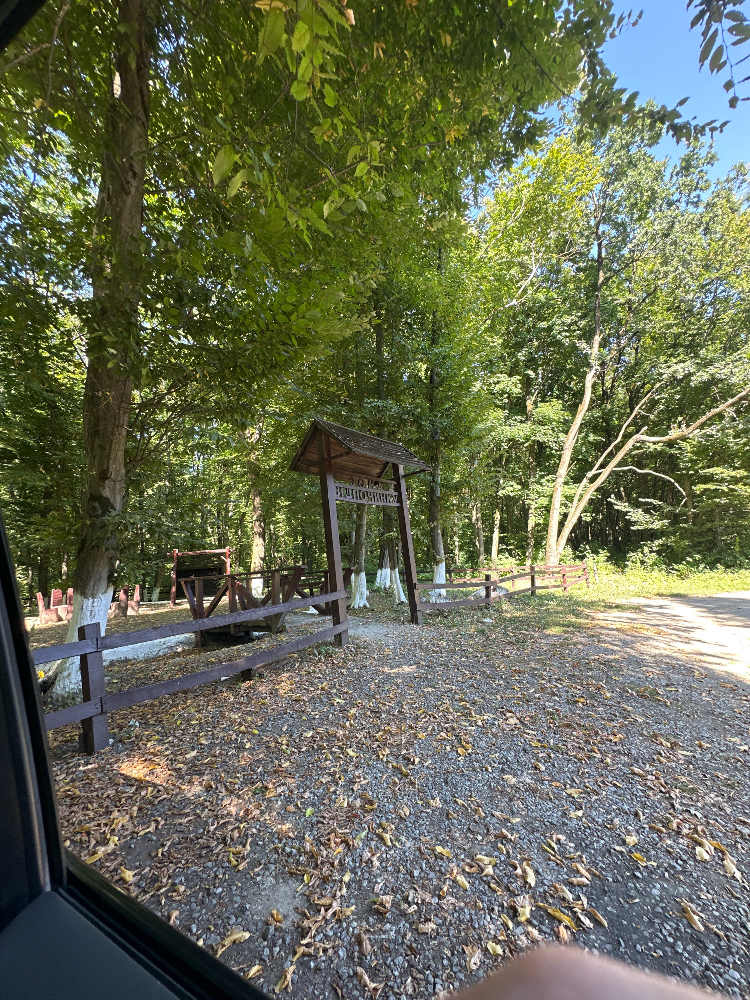 An outdoor scene in Shatava, Ukraine, depicts a wooded area with a small wooden pavilion that has Cyrillic writing on it, possibly indicating a resting spot or attraction. A dark wooden fence encloses an area with picnic tables or benches, and some trees are painted white at their bases. The ground is covered with gravel and fallen leaves, suggesting an autumn setting. A dirt road runs along the right side of the image. The perspective is from a vehicle, with a portion of the car window visible in the foreground. The sky is blue with some clouds, and the sun is shining, casting shadows.