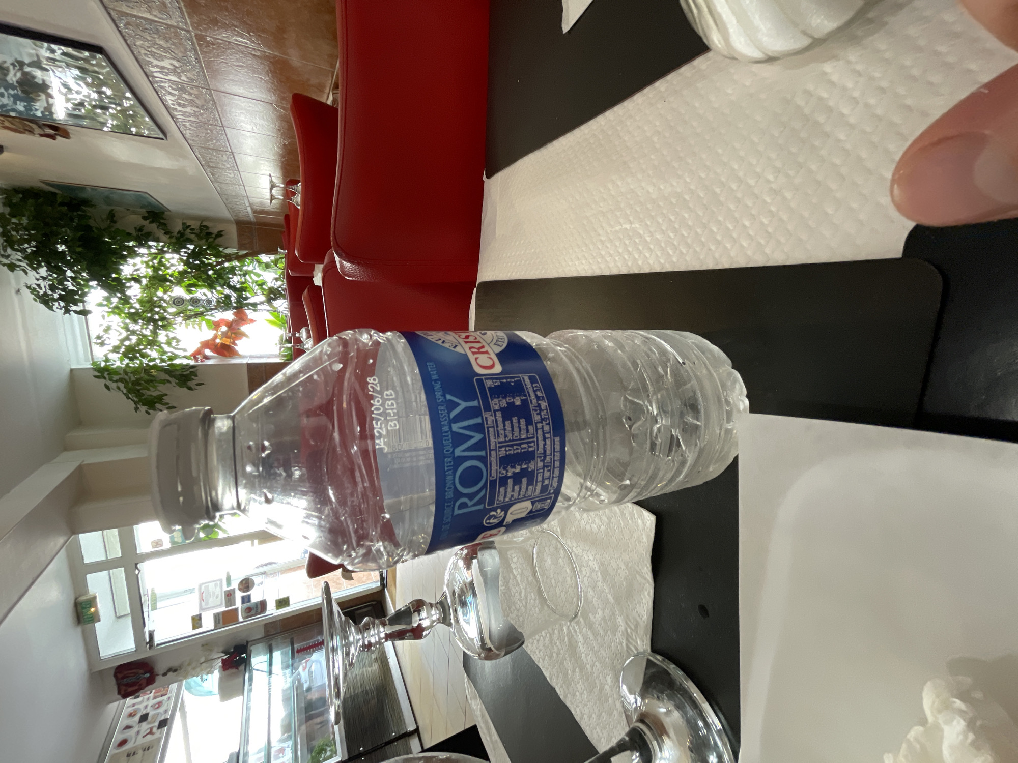The image depicts an indoor scene, possibly a restaurant, with red seating booths visible. On a dark table, a plastic bottle of ROMY brand spring water is prominently featured. A partially visible wine glass sits to the left of the water bottle, placed on a white paper napkin. The background shows a window with abundant greenery, and to the left, a display case with items inside. The overall impression is of a casual dining setting, with the water bottle and glassware suggesting a meal is either in progress or about to begin. The label on the water bottle includes French text, indicating the origin of the product. The scene does not provide any explicit visual cues to pinpoint the exact location in Meaux, France, beyond the general impression of an establishment within that city.
