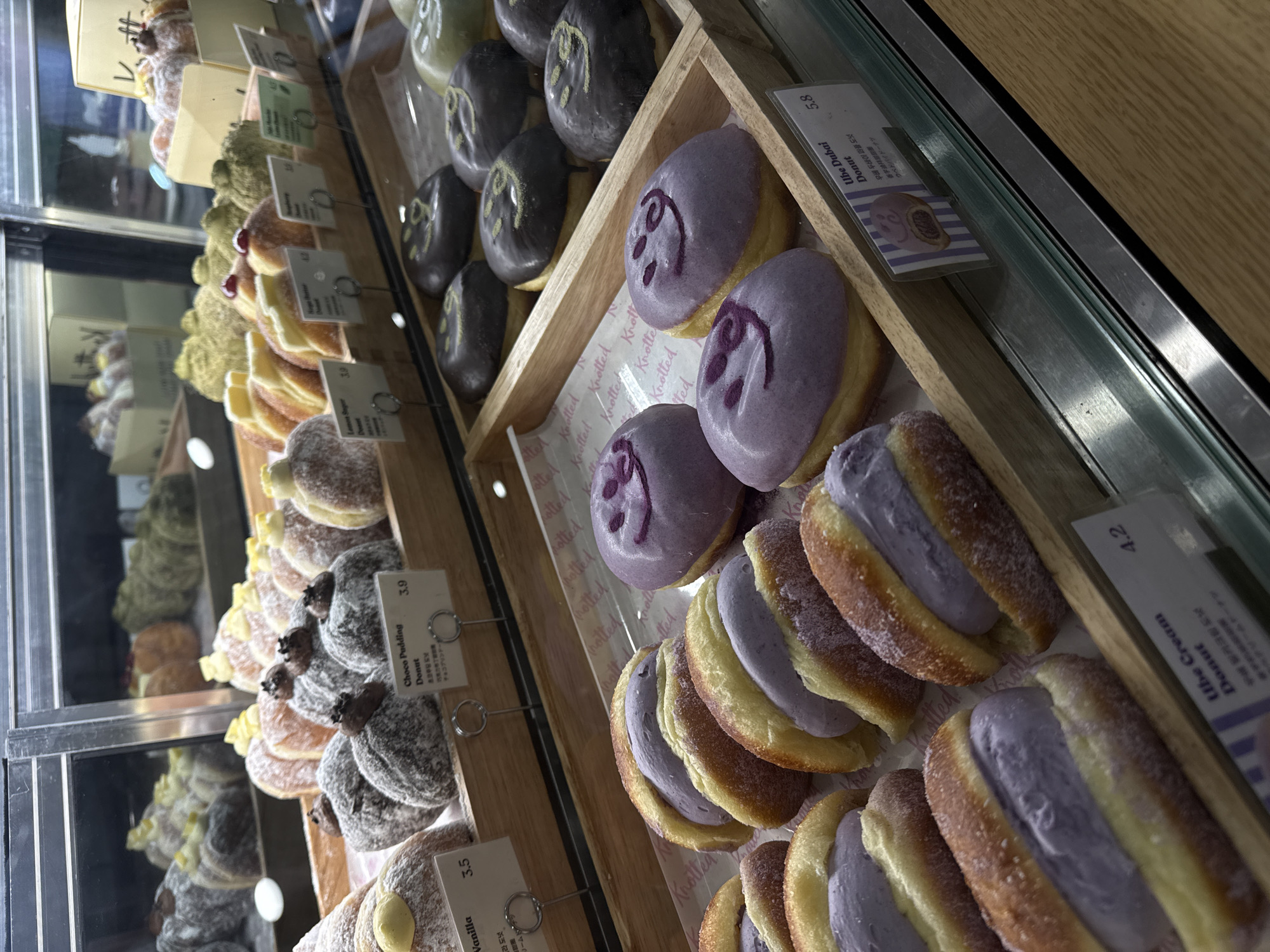 The image captures a selection of doughnuts displayed in a glass-fronted cabinet, likely within a bakery or café located in Gimpo-si, South Korea. The doughnuts are neatly arranged in wooden trays. A variety of flavors are visible, including purple-glazed doughnuts with a face design, "Ube Cream" donuts filled with purple cream and dusted with sugar, chocolate pudding doughnuts, and vanilla doughnuts. Price tags with descriptions in Korean, English, and Japanese are placed near each type of doughnut. The "Ube Dubai Donut" is priced at 5.8. The lighting is bright, highlighting the details and textures of the baked goods.