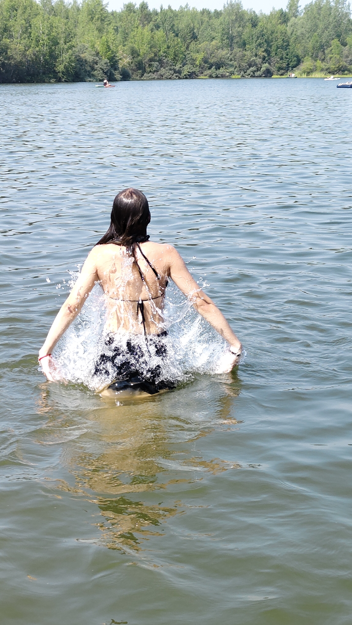 In the city of Terent'yevo, Russia, a person wearing a black bikini is standing in a lake, creating a splash of water. The person's back is to the camera, with their arms outstretched. The water is clear and calm, with ripples extending outwards from the person. In the background, a line of green trees borders the lake, and the sky is clear and blue. The image captures a moment of enjoyment and activity in a natural setting.