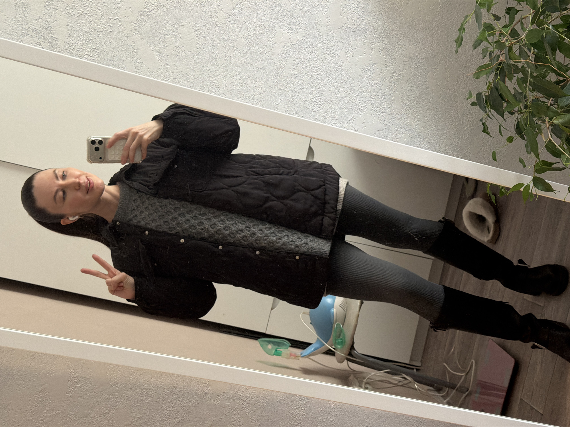 A woman is depicted taking a mirror selfie indoors, in what appears to be a home in Perm, Russia. She stands full-body in the reflection, dressed in a black quilted jacket worn over a grey patterned sweater, grey ribbed leggings, and tall black boots with buckles. Her dark hair is pulled back, and she has small white earbuds. She holds a light-colored smartphone in her right hand, positioned to capture her reflection, while her left hand is raised in a peace sign. Her expression is calm and she has a slight smile.
The background visible in the mirror shows a textured white wall and a wooden floor. To the right, a large green leafy plant is prominent. Near the floor, an off-white fluffy slipper and a medical device, likely a nebulizer with its mask and tubing, are visible. Further left on the floor, there is a white object resembling a humidifier or air purifier, some loose wires, and a small pink/red item. The overall scene suggests a casual moment captured within a personal living space, with the woman's warm attire indicating a cooler climate. No discernible text is visible anywhere in the image.