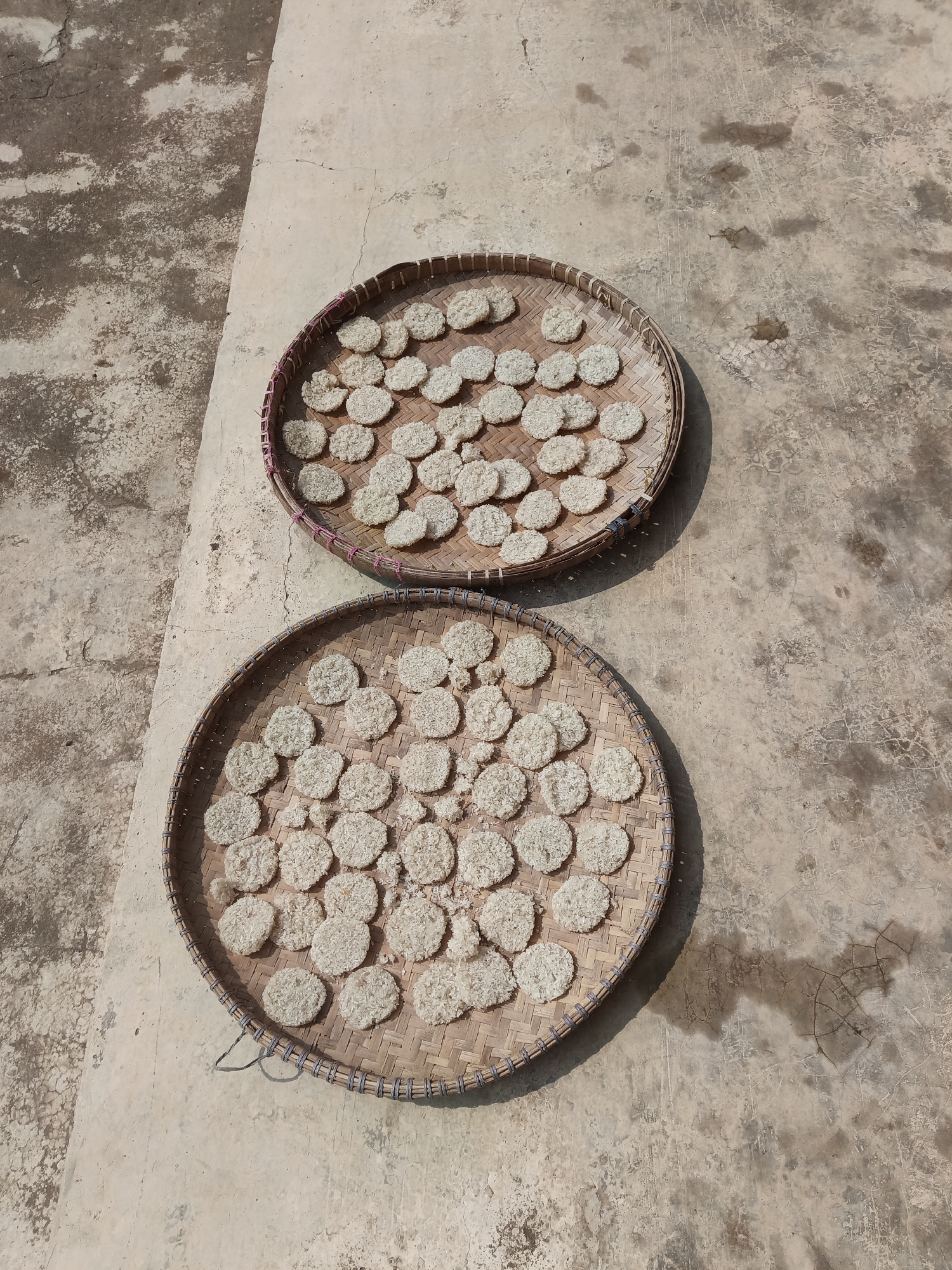 Two round woven bamboo trays, each filled with numerous small, flat, circular food items, are placed on a concrete surface. The food items appear to be rice crackers or similar dried snacks. The concrete surface shows areas of discoloration and cracking, suggesting an outdoor environment. The trays are positioned on the ground, with one slightly overlapping the other. The lighting indicates a bright, sunny day, casting shadows from the trays. The scene is likely a preparation or drying process for food, possibly in a rural or semi-urban setting. This image was taken in Terbanggi Marga, Indonesia.