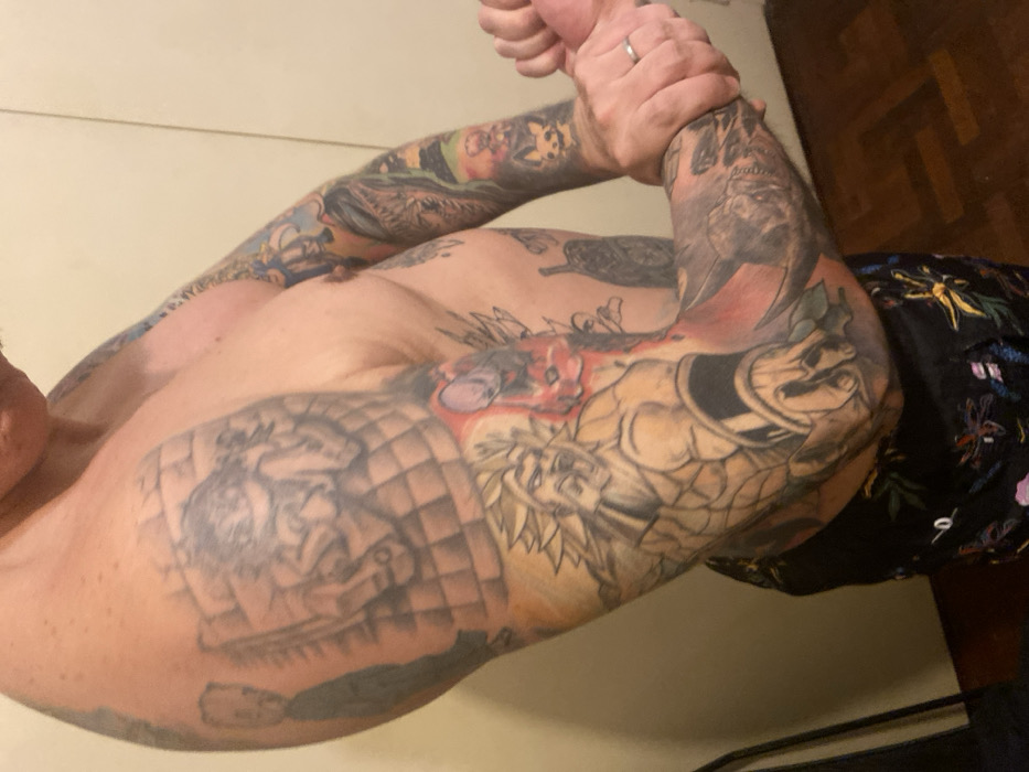 A heavily tattooed, shirtless person poses with arms clasped. Extensive ink covers both arms and chest. The right arm features Zeus with a laurel wreath and a reptile, potentially a turtle, wearing headgear. The left chest displays a shadowed, grayscale image possibly depicting a person within a grid-like structure. The tattoo on the left arm includes cartoon characters. A wedding band is visible on the hand. Part of a black floral shirt is seen on the right side.