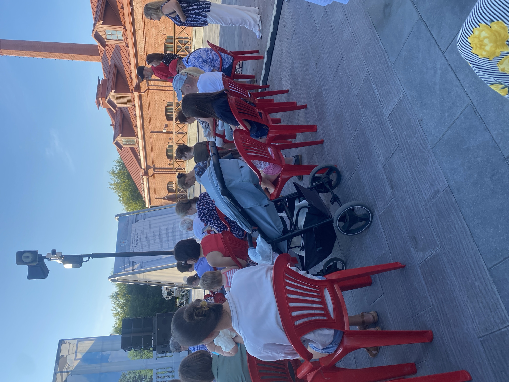 A group of people are seated on red plastic chairs outside, facing a stage area. The setting appears to be an outdoor event, possibly a performance or lecture, given the stage setup with speakers and a large display board. The architecture visible in the background, characterized by red brick buildings with arched windows and dormers, suggests a historical or cultural district. The presence of a baby stroller indicates that families are attending the event. The lighting suggests it is daytime, with a clear blue sky. The flooring is paved with large, rectangular tiles. The overall scene is that of a public gathering in an urban environment. Without specific landmarks or textual clues within the image, it is not possible to pinpoint the exact location in the city of Minsk, Belarus.