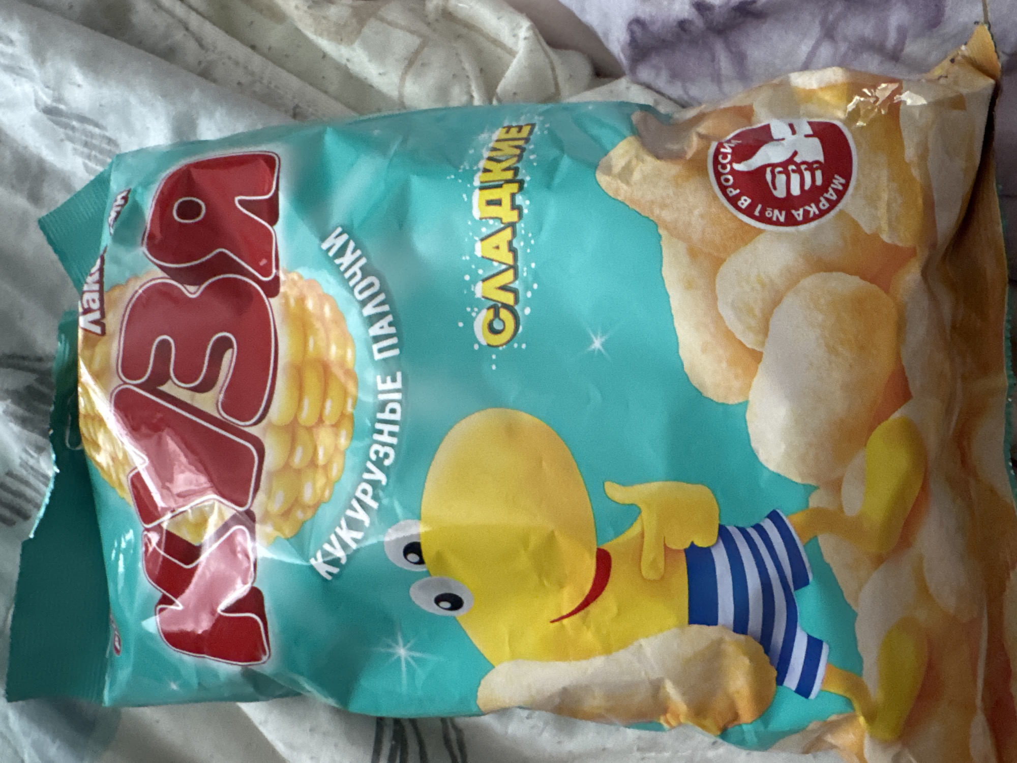 The image is a close-up, eye-level photograph of a bag of "Кузя" brand corn snacks. The bag is primarily teal-colored with the brand name "Кузя" in large red lettering across the top. Below the brand name, smaller white text reads "КУКУРУЗНЫЕ ПАЛОЧКИ" (corn sticks). A cartoon yellow character with large eyes and a red smile, wearing blue and white striped shorts, is depicted on the left side of the bag. To the right of the character, the word "СЛАДКИЕ" (sweet) is written in yellow. A red circular logo with a white thumb-up icon and text "МАРКА №1 В РОССИИ" (Brand #1 in Russia) is located in the lower right quadrant of the bag. The bag is resting on a surface that appears to be a light-colored, possibly white, fabric, likely a bedsheet or blanket. The lighting suggests an indoor environment. There is no indication of the city of Saratov, Russia, or any specific urban location within the image.