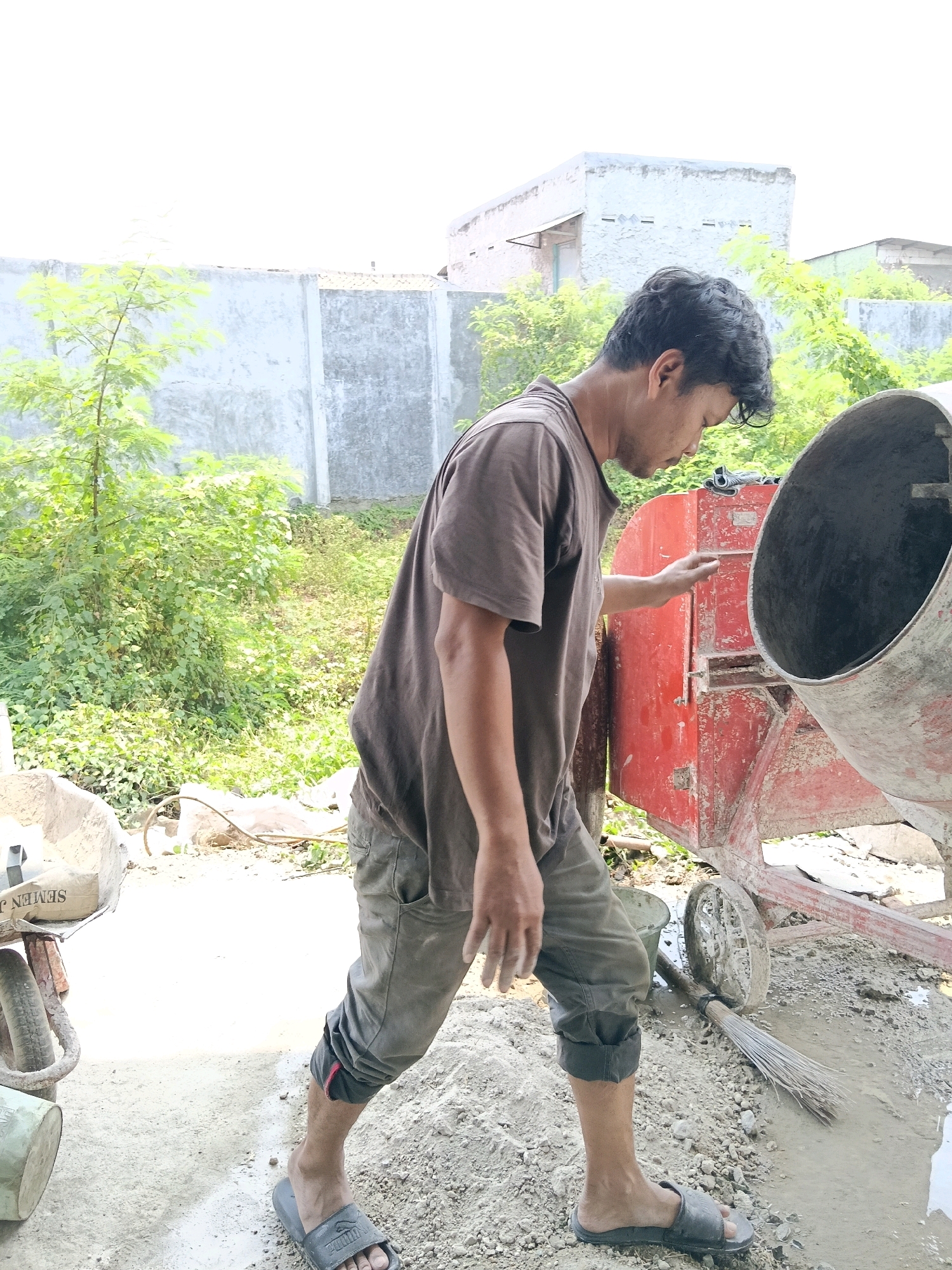In the city of Belimbing, Indonesia, a construction worker is seen from the side near a cement mixer. The man, wearing a brown t-shirt and grey cropped pants, is looking down at the mixer with his right hand raised as if interacting with it. A wheelbarrow with a bag of cement is to his left, and a broom lies on the ground to his right. The background features a concrete wall and lush green foliage, suggesting an outdoor construction site. The ground is covered in sand and some wet cement.