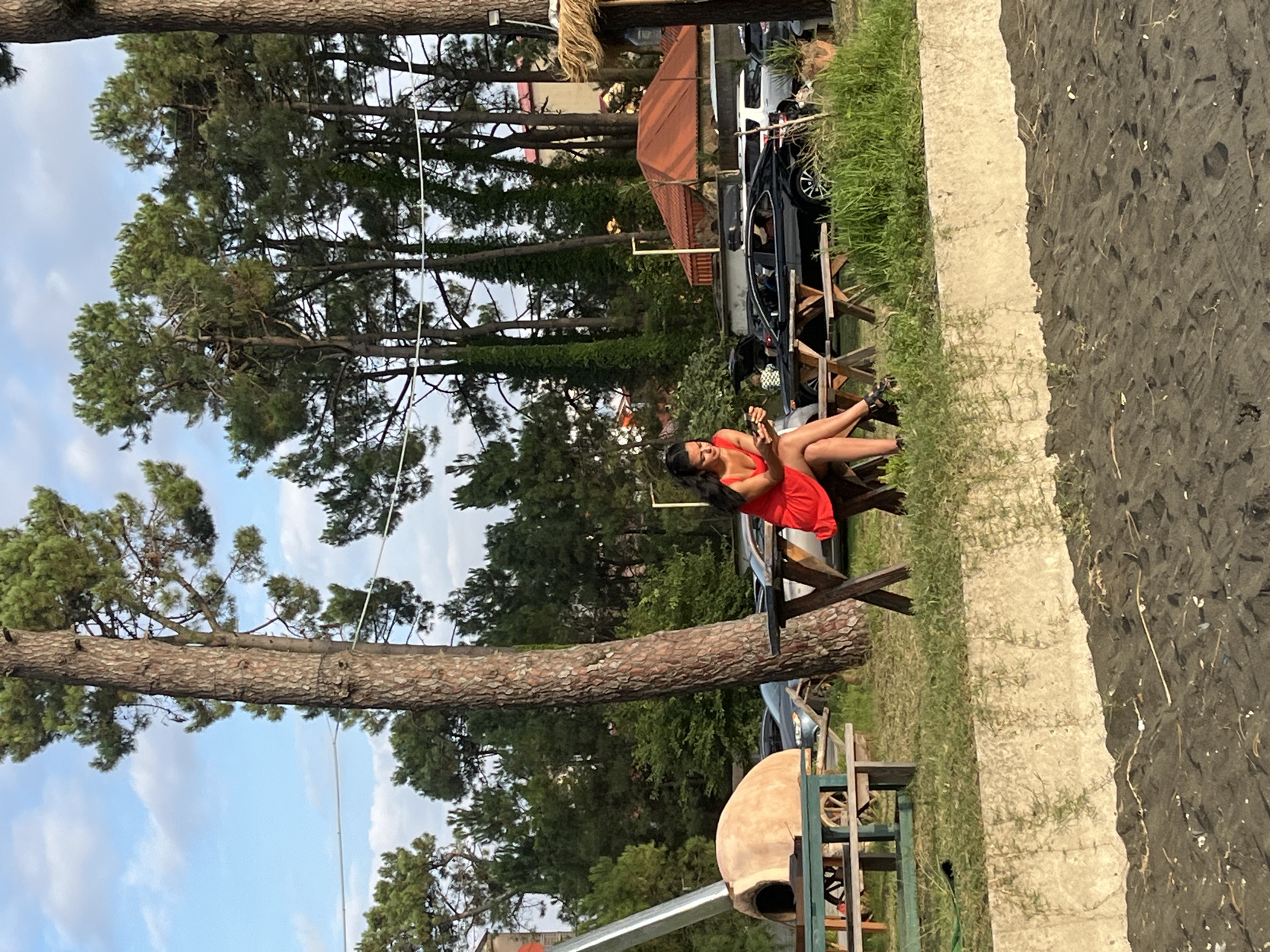 A woman is sitting at a picnic table in Ureki, Georgia, looking at her phone. She is wearing a red dress and black shoes. She is sitting on a wooden bench at a black table with X-shaped legs. There are trees in the background, as well as cars and buildings. The ground in front of the table is covered in grass and a concrete wall, with black sand in the foreground. There is a wood-fired oven in the background to the left.