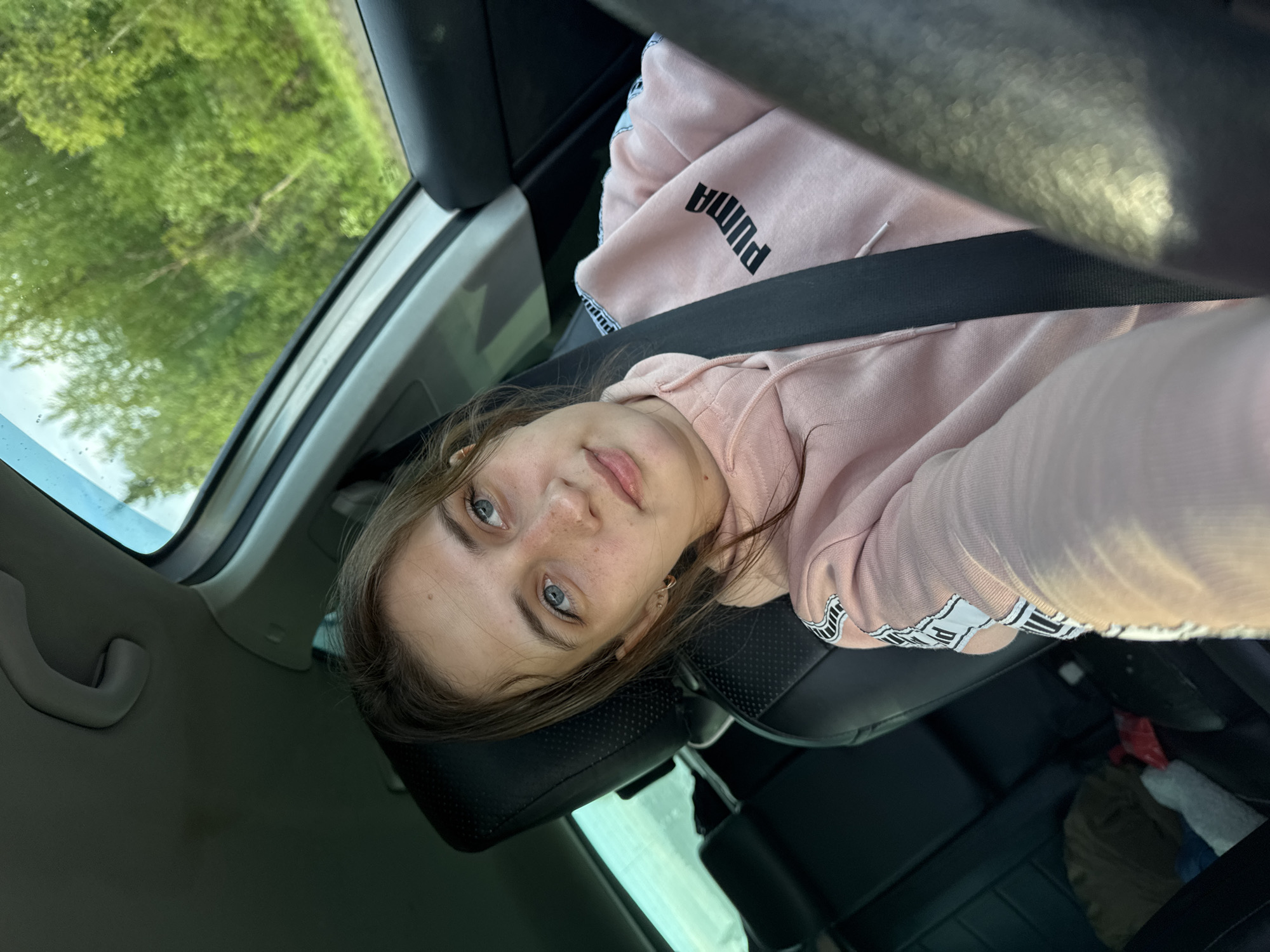 A young woman with blue eyes and freckles is seated in the front passenger seat of a car. She is wearing a pink Puma hoodie and a seatbelt. The background visible through the car windows shows green trees, which could be indicative of a location near Malakhovo, Russia, given its proximity to forested areas. The interior of the car has black leather seats.