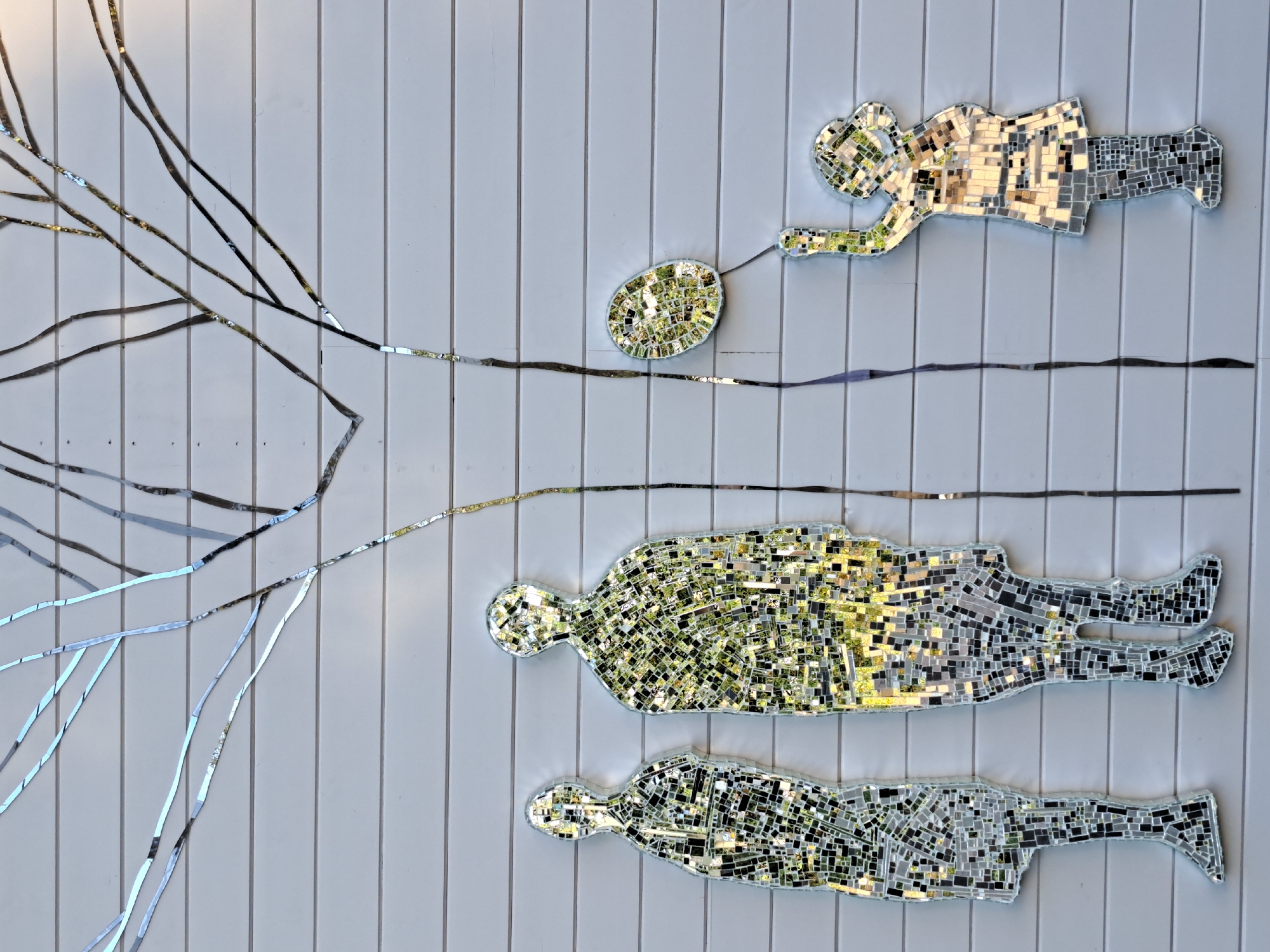 The image depicts a mosaic artwork on a horizontally planked, light-colored wall. The mosaic features stylized human figures and tree branches, all rendered with mirrored tiles that reflect the surrounding environment.
On the left, two tall, slender human figures stand side-by-side. The figure on the left is slightly shorter and thinner than the one on the right. Both figures are composed of numerous small, irregular mirrored pieces, creating a fragmented, shimmering effect. The reflections within the tiles suggest greenery and possibly the sky.
To the right of the two figures is a mosaic of a young child. This figure is depicted holding a balloon above its head. The child is also made of mirrored tiles, reflecting a similar palette of colors to the adult figures. A thin, dark line, also appearing to be made of mosaic pieces, extends vertically from the upper right of the frame down towards the child, possibly representing a tree trunk or branch.
Above the figures, the mosaic extends to form a representation of tree branches. These branches are created from thin, elongated strips of mirrored material, creating a delicate, branching pattern that spreads across the upper portion of the wall. The reflections in these branch pieces are more elongated and abstract.
The wall itself is a uniform, light color, with distinct horizontal lines indicating the planks of its construction. The lighting suggests it is daytime, with the sun casting highlights and shadows on the mosaic pieces, enhancing their reflective qualities.
This artwork is located on the facade of the Irkutsk Regional Art Museum in Irkutsk, Russia. The specific artwork is titled "Reflection" by Alexander Smirnitsky.