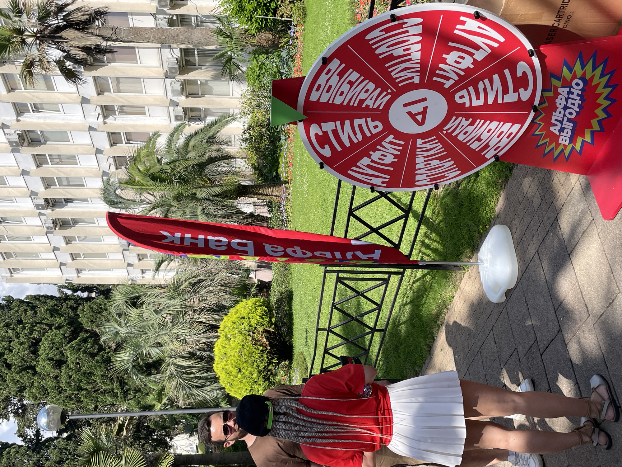 The image captures a scene in Sochi, Russia, likely near a public building or park. In the foreground, a spinning prize wheel, predominantly red with white lettering, is prominently displayed. The wheel has several segments with Russian text. Below the wheel, a blue and yellow starburst graphic features the text "АЛЬФА ВЫГОДНО," which translates to "Alfa is profitable."
To the left of the wheel, two individuals are visible. A man in a brown t-shirt and sunglasses stands facing away from the camera, partially obscured by a woman. The woman, with distinctive long, braided grey hair adorned with a green band, is facing the wheel. She is wearing a red t-shirt and a short white pleated skirt.
A tall, red banner with white Cyrillic lettering, "Альфа Банк," stands behind the woman and to the left of the wheel. In the background, a lush green lawn is dotted with flowers, and a wrought-iron fence runs horizontally. Beyond the lawn, a modern building with multiple windows and air conditioning units stands under a partly cloudy sky. Palm trees and other dense foliage are scattered throughout the scene, typical of a subtropical environment like Sochi. A lamp post with a spherical fixture is visible on the far left.