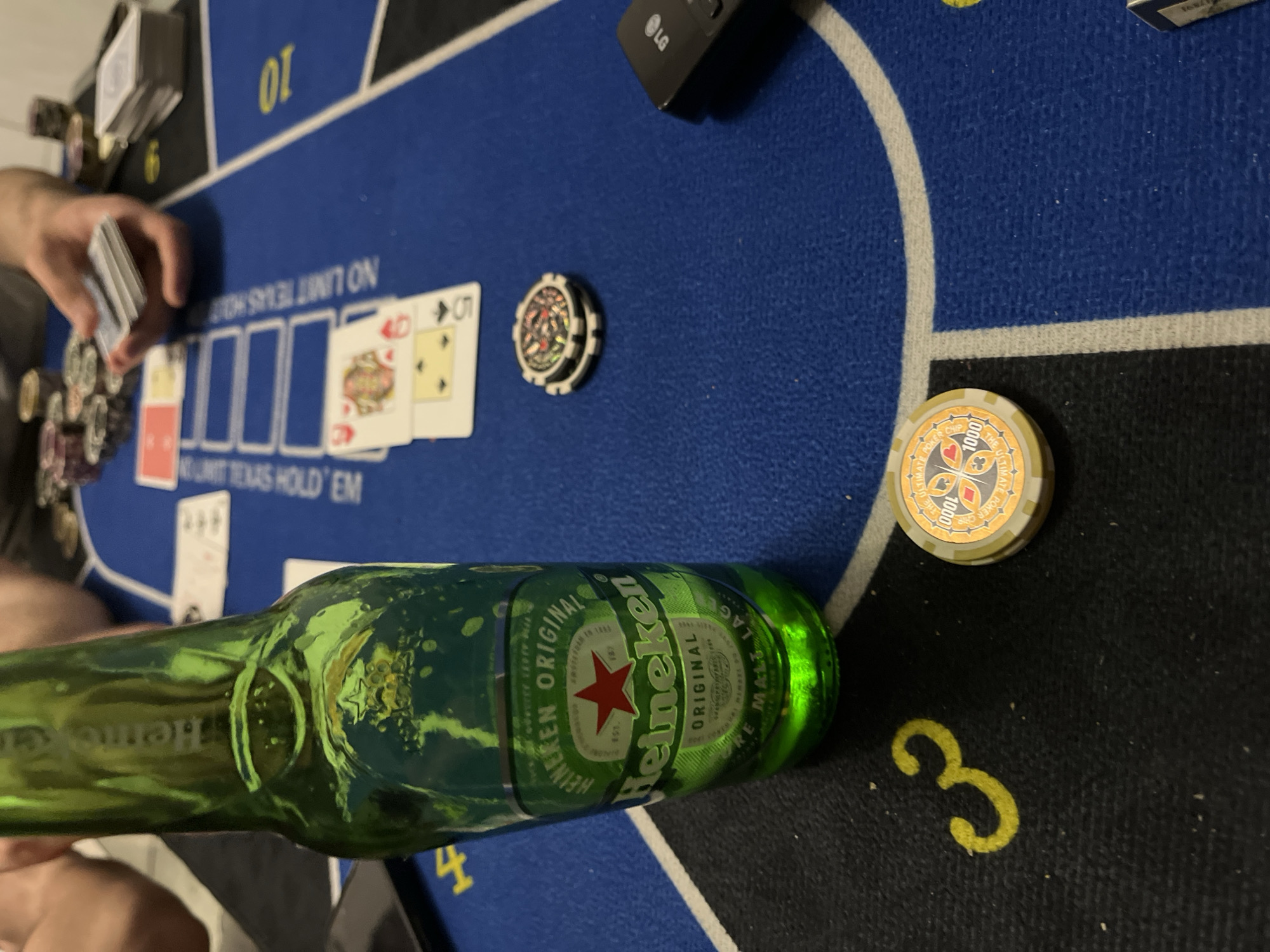 The image depicts a poker game in progress. A person is holding a deck of cards while other cards (5 of clubs and 4 of diamonds) and chips are visible on the table. There is a Heineken beer bottle laid on its side next to more chips, and an LG remote. The poker table itself is patterned with numbered sections and reads "NO LIMIT TEXAS HOLD 'EM". The location is not discernable from the image. There is no information about the city of Burgas, Bulgaria.