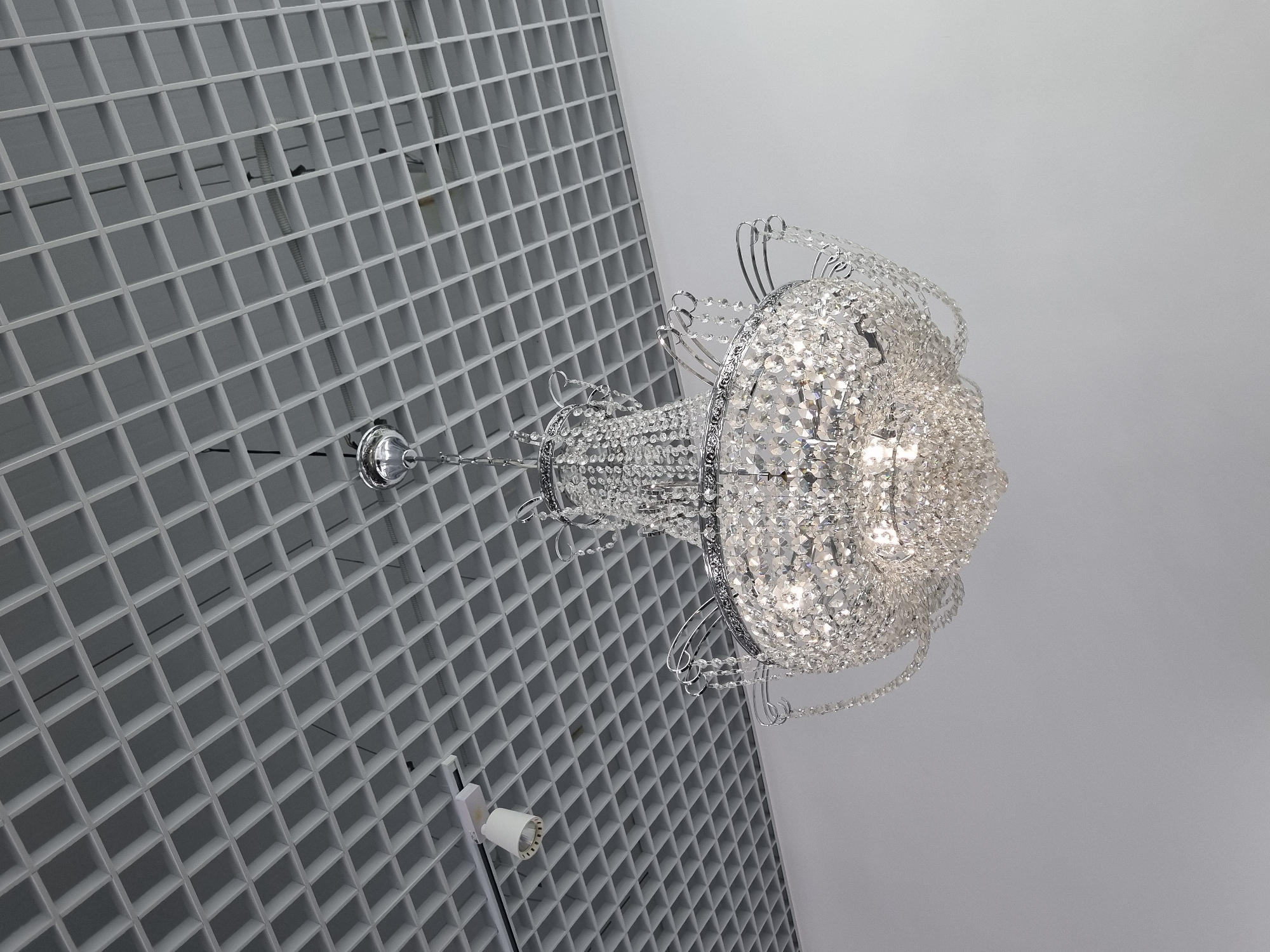 An overhead view depicts a large, illuminated multi-tiered crystal chandelier suspended from a white open-cell grid ceiling. The chandelier features a polished chrome or silver-colored frame adorned with numerous faceted clear crystal elements, catching and refracting light from internal sources. It hangs by a single chrome chain and canopy. The grid ceiling, likely a 'Grilyato' system, reveals darker structural elements and utilities above. To the upper left, a white track-mounted cylindrical spotlight is visible, directed downwards. The walls below the ceiling appear uniformly light-colored. No individuals are present. The specific location within the city of Chaikovsky, Russia, cannot be determined from the visual evidence provided. The environment suggests an indoor commercial or public space.