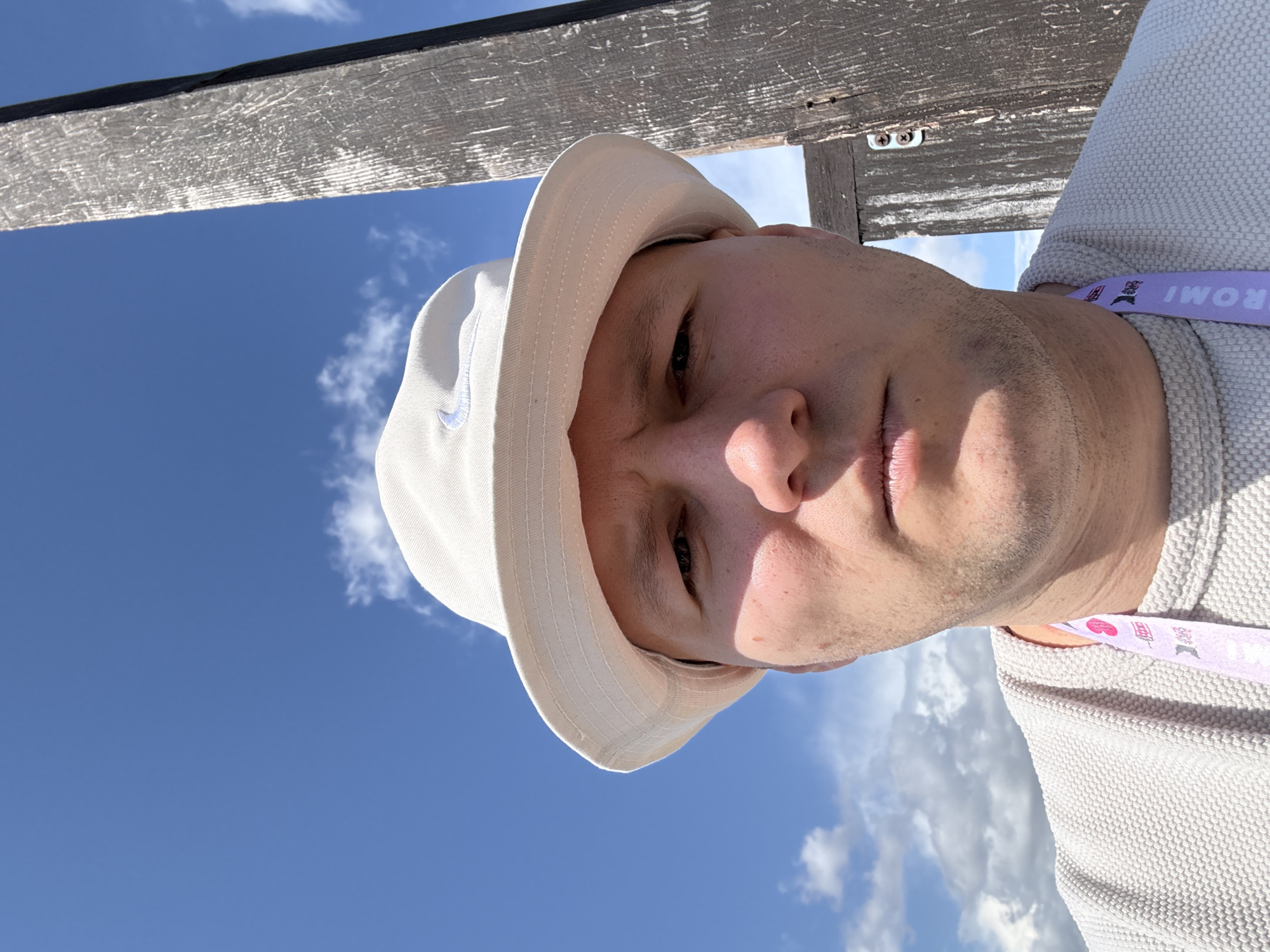 A man is pictured in an upward-facing, close-up selfie. He is wearing a light-colored bucket hat with a visible swoosh logo. He also wears a light-colored textured shirt and a lanyard around his neck. Above him, a wooden structure is partially visible against a backdrop of a blue sky with scattered white clouds. The location of the picture is Старцево, Russia.