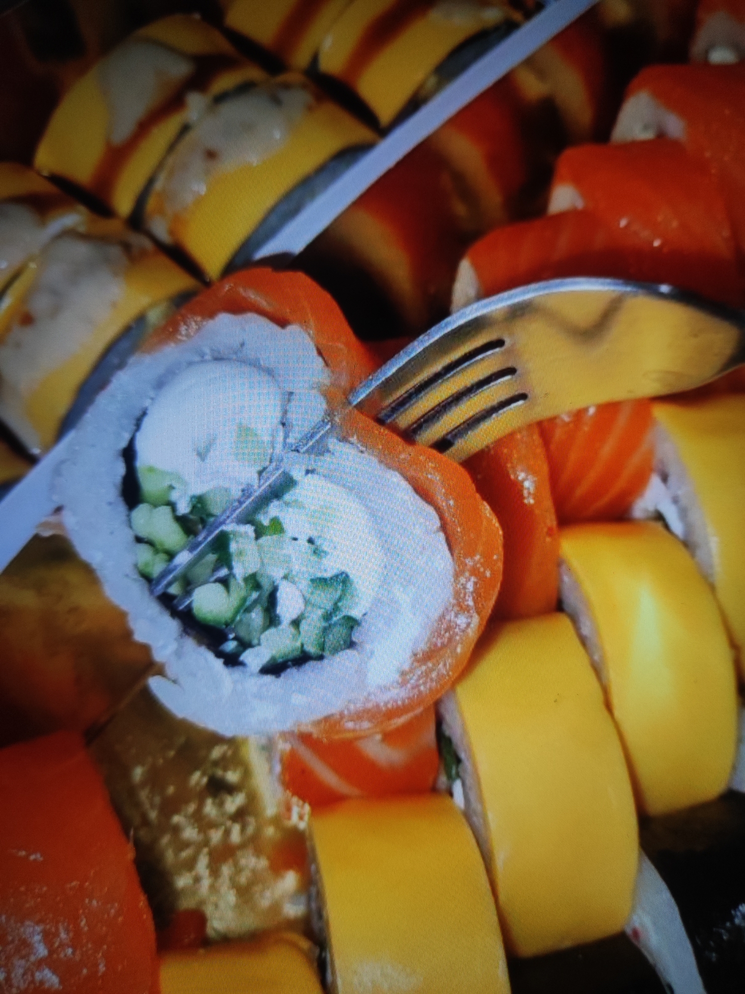 The image is a close-up, top-down view of a plate of sushi rolls. A fork is positioned over the sushi, with its tines digging into one of the rolls. The sushi rolls are various colors, including orange (likely salmon), yellow (possibly mango or egg), and white (rice). The interior of the roll being pierced by the fork appears to contain green cucumber and white cream cheese. The background shows more sushi rolls in a similar arrangement, suggesting they are part of a larger platter or serving. The lighting is bright, causing some glare on the sushi and fork. There is no indication of a specific location in Cherkasy, Ukraine.