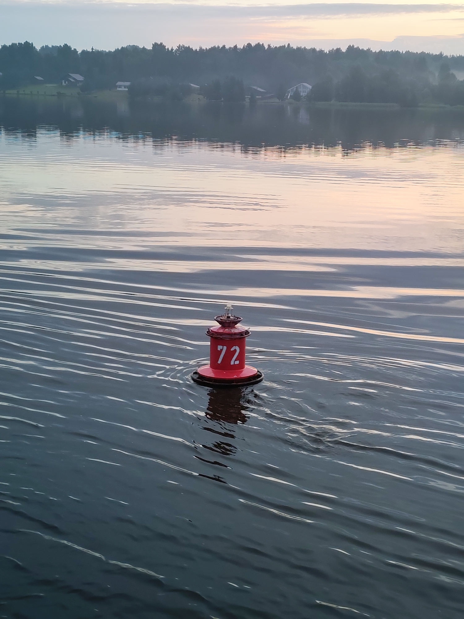 A red buoy with the number "72" in white is floating on the dark, rippling water of a lake or river. The water's surface reflects the soft, pastel colors of the sky at dawn or dusk, with streaks of pale pink and blue. In the background, a hazy treeline is visible, dotted with a few houses, suggesting a rural or suburban setting. The overall atmosphere is serene and tranquil. This scene is located in Kondratovo, Russia.