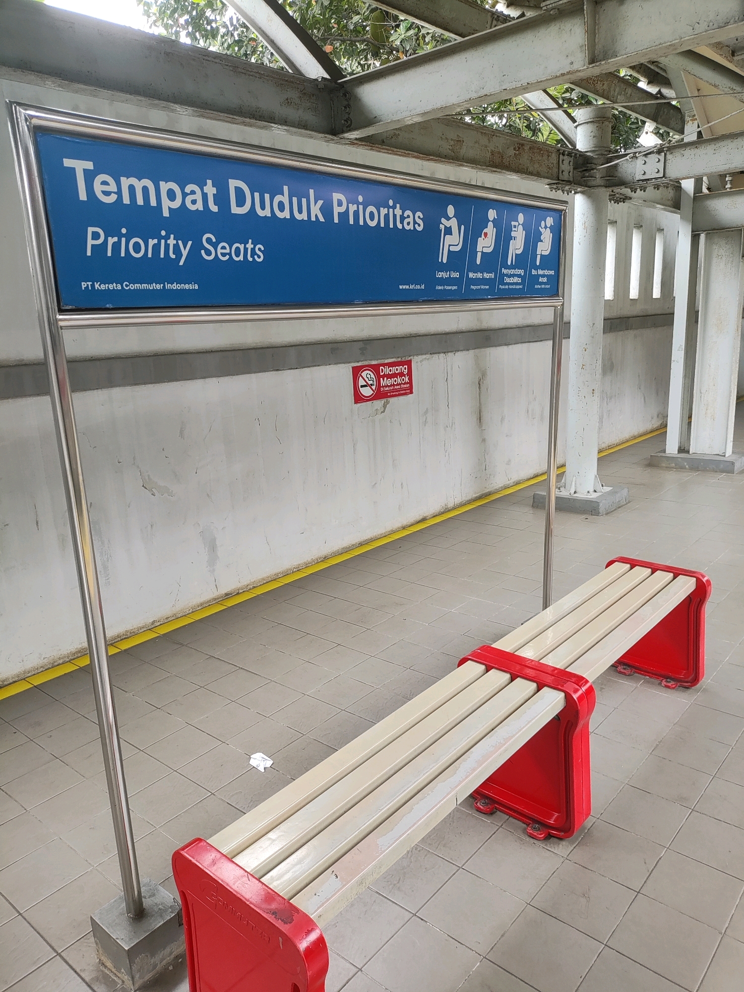 A long shot captures a platform at the Telagamurni, Indonesia train station. A blue sign with white text reads "Tempat Duduk Prioritas" and "Priority Seats" in English, accompanied by pictograms of individuals representing elderly passengers, pregnant women, passengers with disabilities, and mothers with children. Below this sign, a red and white bench is positioned for seating. To the left of the bench, a metal pole anchors the sign. A "Dilarang Merokok" (No Smoking) sign is visible on the wall behind the bench. The platform is paved with grey tiles, with a yellow line marking the edge. The area is partially covered by an overhead structure with metal beams. No people are present in the image.