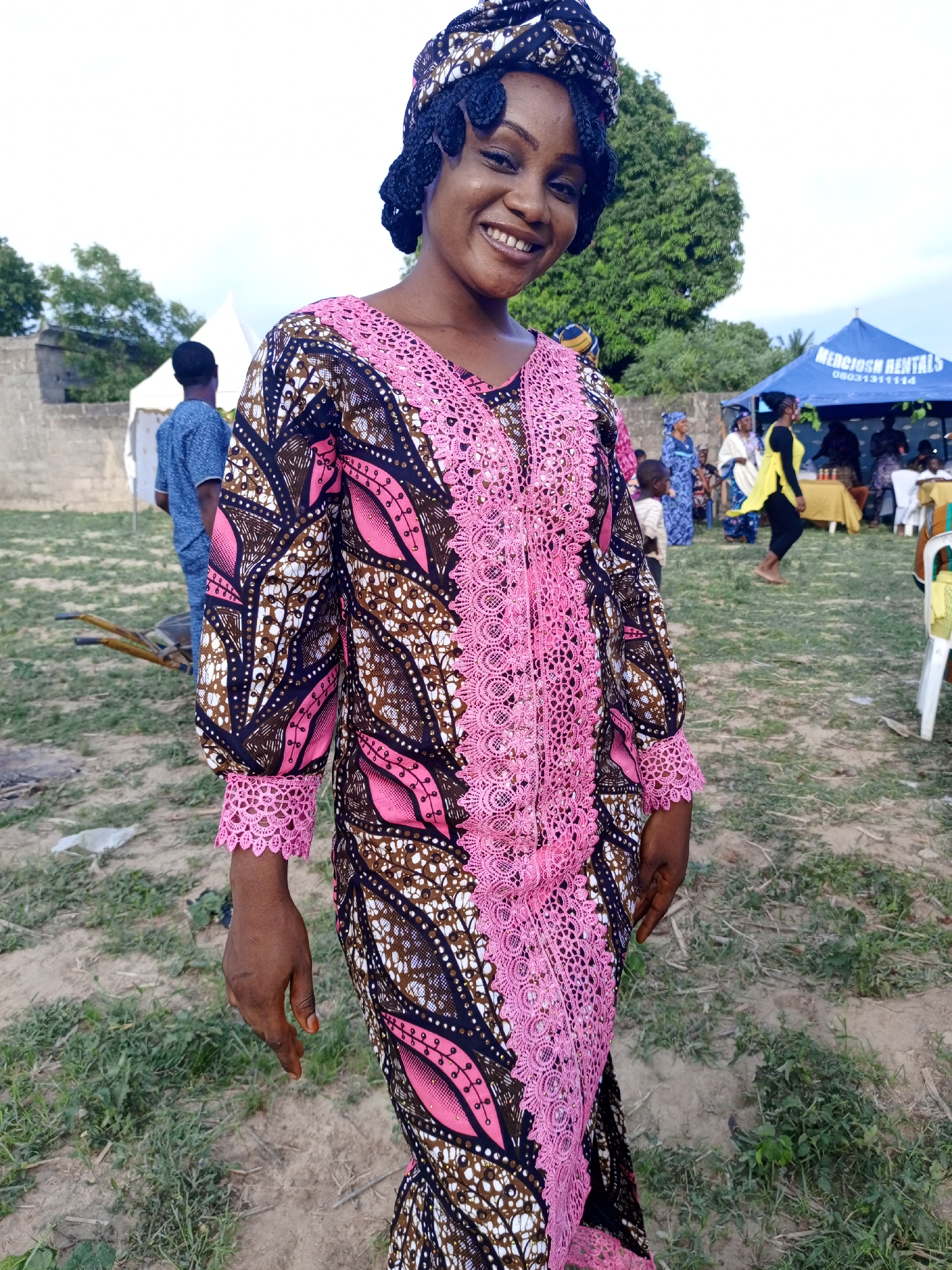 The image shows a smiling woman wearing a patterned dress with pink lace trim. She has her hair wrapped in a headscarf. The location appears to be an outdoor event on a grassy area, possibly in Keffi, Nigeria, given the prompt's mention. People are visible in the background, along with a tent and some chairs, suggesting a gathering or celebration. A partial view of a building or wall is also seen in the background.