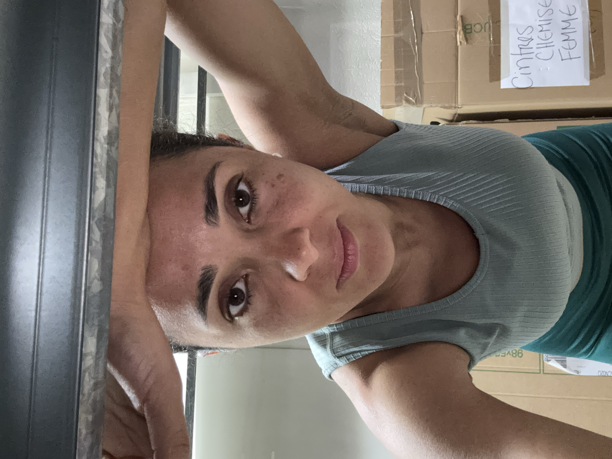 A woman is pictured indoors, with boxes stacked in the background. The boxes appear to be labeled in French, one reading "Cintres : Chemise Femme." The location cannot be determined to be Luxembourg City, Luxembourg, based on the visual information alone.