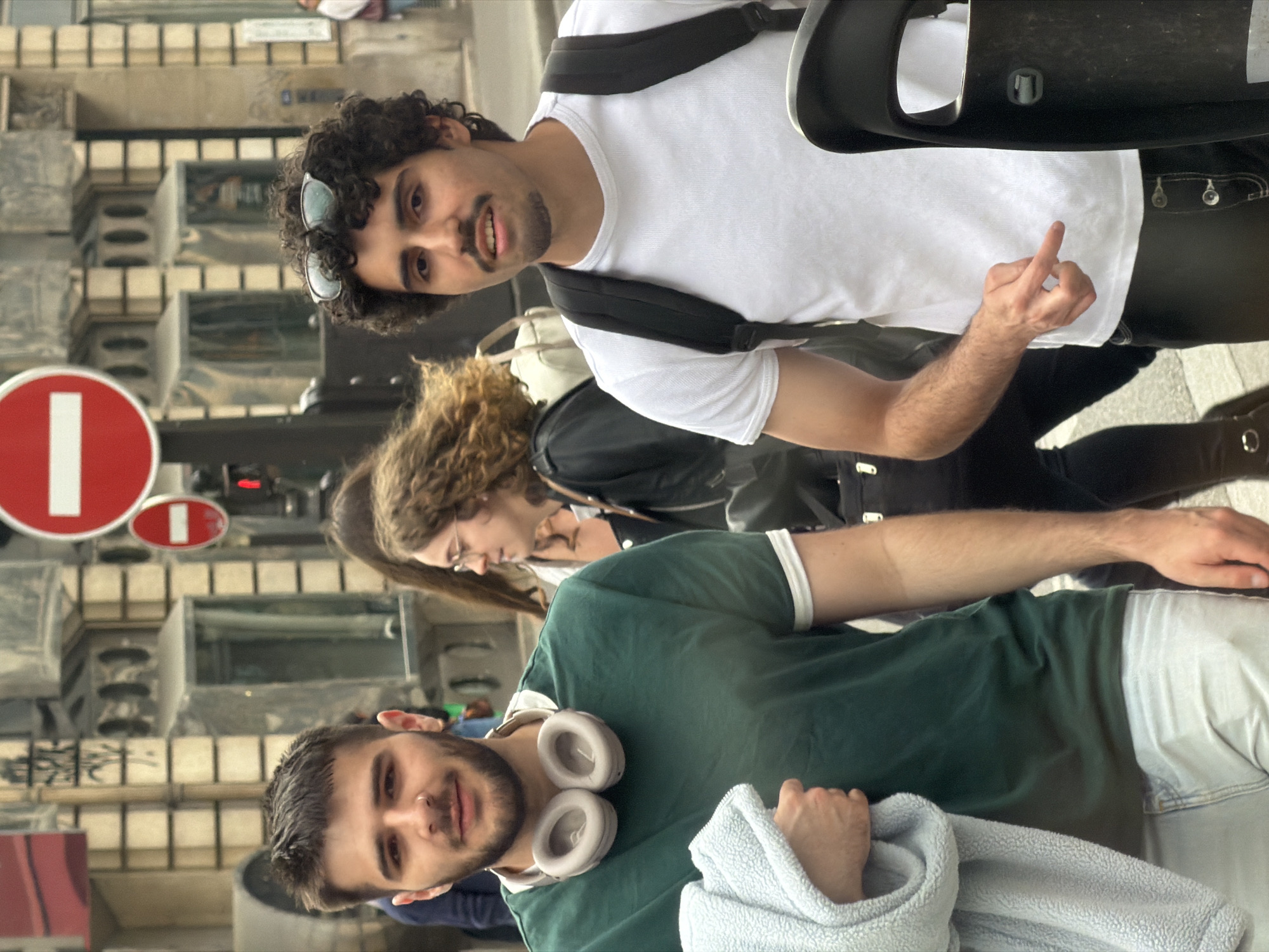 The image captures an outdoor scene in Paris, France, with a group of four people. A man in a green polo shirt and light-wash jeans is in the foreground, wearing white headphones around his neck and holding a light blue towel. Behind him is a woman with curly hair wearing glasses and a black jacket. Next to her is a man in a white t-shirt, black pants, and a black backpack, gesturing with his middle finger raised. The background features a Parisian building with a distinctive facade and "No Entry" road signs. The photo appears to be candid, capturing a moment in the urban environment.