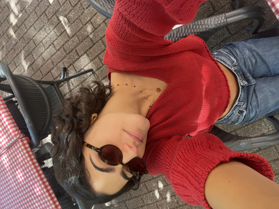 A young adult female is depicted in a high-angle selfie, seated outdoors on a patio in Orléans, France. She has dark, curly hair and wears tortoise-shell framed oval sunglasses, a red V-neck cropped knit sweater, and blue denim jeans. Her visible right ear features a gold stud earring, and she wears a double-layered gold necklace with small red beads and a heart-shaped pendant. The background shows dark, woven-style outdoor chairs and a table covered with a red and white checkered tablecloth. The ground is paved with light-colored rectangular bricks, illuminated by natural sunlight casting shadows.