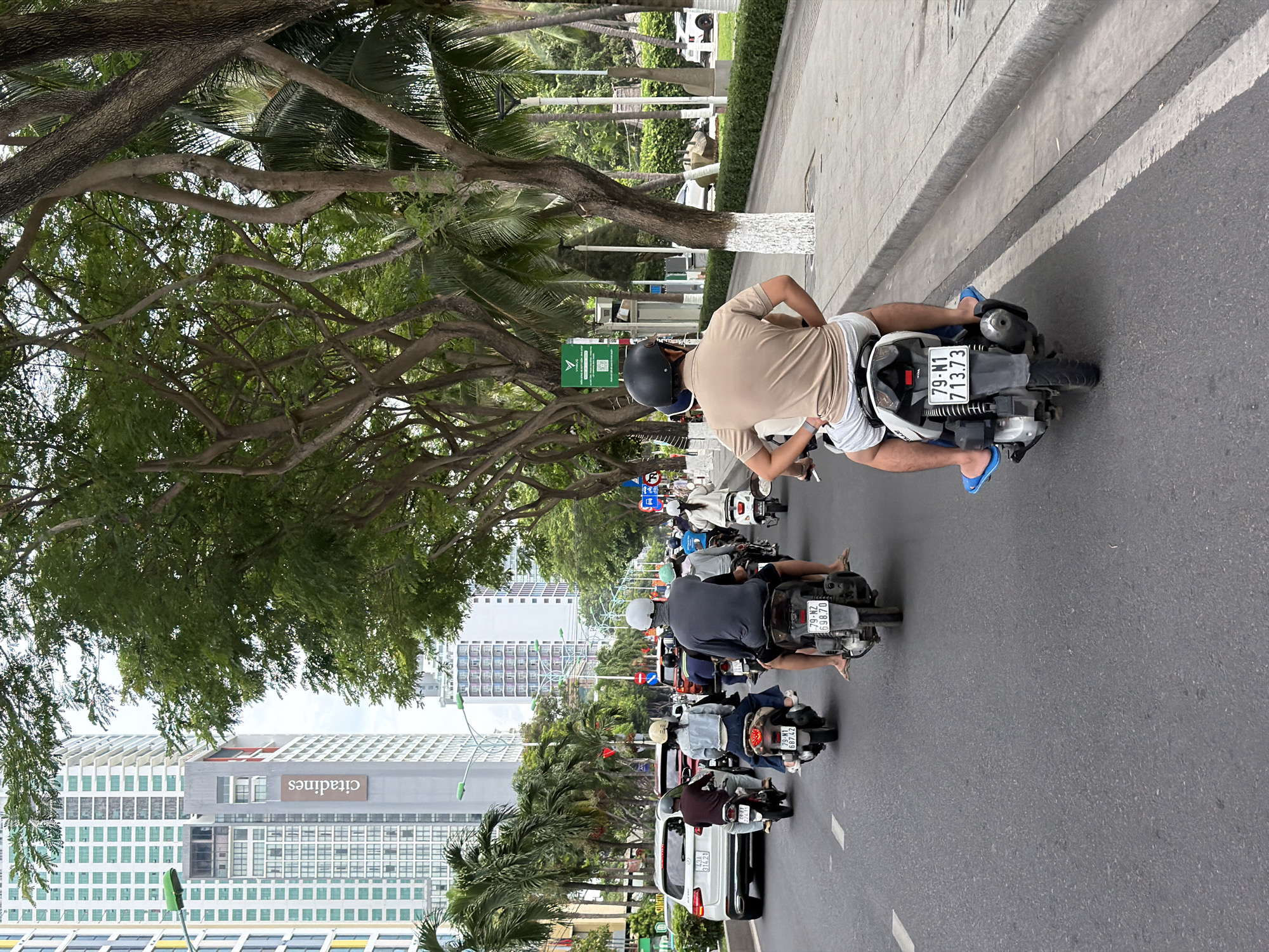 This image captures a street scene in Nha Trang, Vietnam, likely along a coastal road given the presence of palm trees and a sidewalk bordering a paved area. Multiple motorbikes are in motion on the asphalt road. In the foreground, a person wearing a tan polo shirt and grey shorts rides a white scooter, their bare feet resting on the blue footrests. The scooter has a license plate that reads "79-N1 713.73".
Behind this scooter, other motorbikes are visible. One rider in a grey shirt and a white helmet is on a black scooter with a license plate "79-NZ 698.70". Further ahead, a rider in a blue denim jacket and white helmet is on a beige scooter. Several other motorbikes and their riders are scattered throughout the middle ground.
The backdrop features tall, modern buildings with numerous windows, characteristic of urban development. Lush green trees, including palm trees, line the street, providing shade. A sidewalk with a white-painted tree trunk and a traffic sign can be seen to the right. The overall impression is a busy street with typical urban traffic in Vietnam.