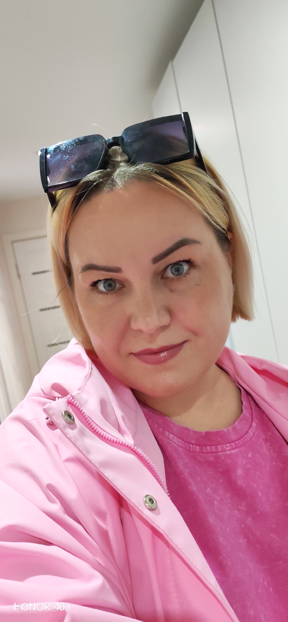 This image depicts a single adult female in an indoor setting within the city of Noviy, Russia. The subject is a fair-skinned individual with blonde, shoulder-length hair, parted centrally. She possesses blue-grey eyes and a subtle, closed-mouth expression, with her lips appearing to have a light gloss. Black, rectangular sunglasses are positioned on the top of her head, with noticeable smudges or reflections on the lenses. She is wearing a bright pink zipped jacket over a darker, possibly stone-washed, pink t-shirt. A metal snap button is visible on the jacket near her right shoulder. The background consists of light-colored walls; a white paneled door is partially visible to the left, and light-colored cabinet or wardrobe panels are present to the right. The lighting is even, suggesting an indoor light source. The image, a close-up from a slightly upward angle, appears to be a selfie, indicated by the "HONOR 400" watermark in the lower left corner.