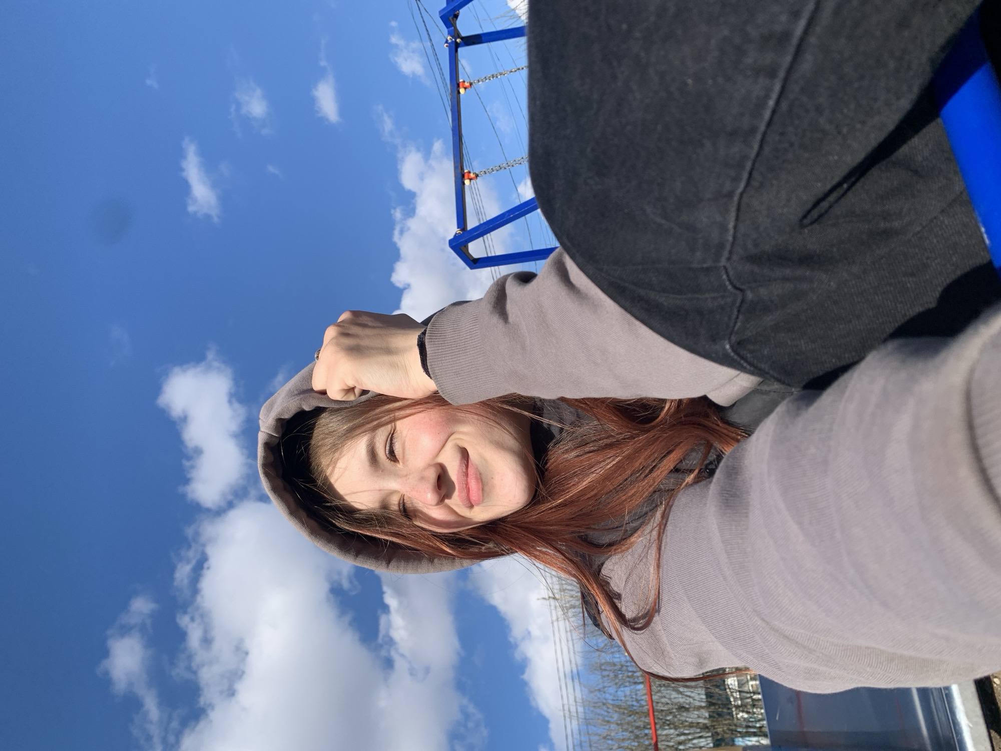 A young person with auburn hair wearing a gray hooded sweatshirt is looking up and smiling. The background shows a blue sky with scattered white clouds and part of a blue playground structure with chains. There are trees with bare branches in the distance, and the weather appears to be sunny. The location is inferred to be in Фоки, Russia.