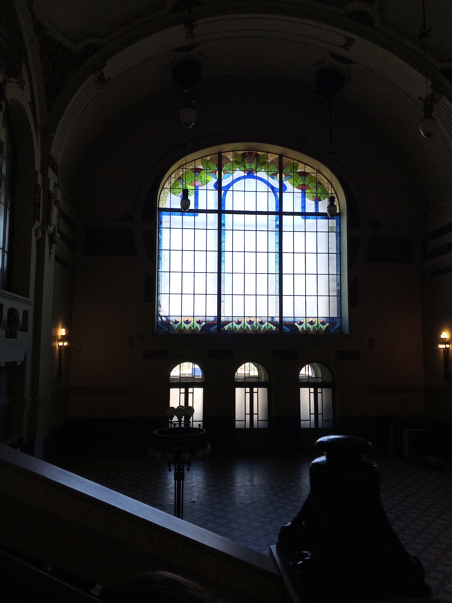 This image depicts the interior of a grand hall within the Vitebsky Railway Station in Saint Petersburg, Russia. The scene is dimly lit, dominated by light entering through a large, arched window structure.
In the center of the background, a prominent, multi-paned arched window is visible. The upper portion of this window features an ornate stained-glass design with a central blue arch bordered by geometric patterns in green, red, and yellow, while the lower main section of the window is clear glass arranged in a grid pattern. Below the large window, three smaller arched openings, appearing to be glass-paneled doorways or windows, are illuminated.
The hall features a high, arched ceiling with visible architectural moldings and two dark, cylindrical pendant lamps suspended. Two wall-mounted sconces on the left and right walls emit a warm, orange glow. In the mid-ground, a dark, decorative floor lamp or candelabra-like fixture stands, its multiple bulbs visible but indistinct. The floor exhibits a dark, possibly tiled or checkered pattern.
In the immediate foreground on the right, a dark, sculpted newel post or baluster is present, partially obscuring the lower-right corner. A dark handrail extends diagonally from the lower-left corner towards the center. No individuals are present, and no actions or interactions are depicted. The overall impression is one of architectural grandeur and historical significance, illuminated by a mix of natural and artificial light sources.
