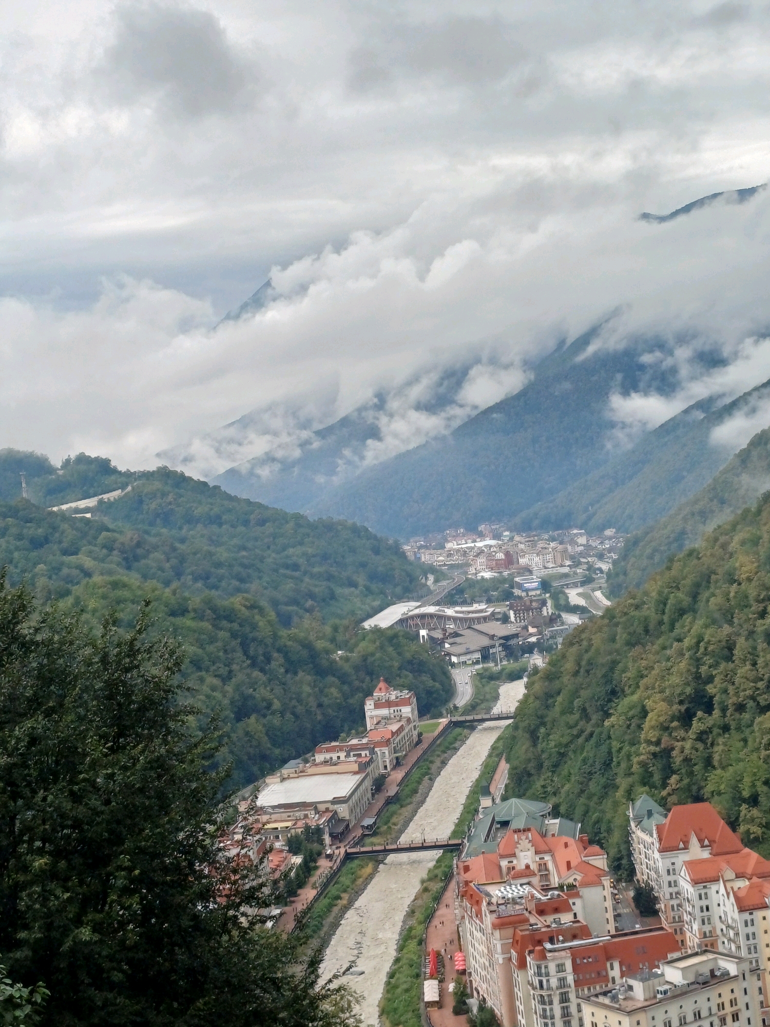 The image captures a wide aerial view of the city of Estosadok, Russia, nestled within a mountainous valley. The sky is overcast with dense, grey clouds partially obscuring the peaks of the surrounding mountains, which are covered in lush green forests. A river, appearing murky and brown, flows through the center of the valley, with a town built along its banks.
The town consists of a dense collection of buildings with red-tiled roofs and white or light-colored facades, suggesting a modern resort or residential area. Roads and infrastructure, including bridges, are visible weaving through the urban landscape. The architecture is a mix of multi-story buildings and smaller structures.
In the foreground, a large, dark green tree with dense foliage partially obstructs the view of the town. The overall impression is one of a settlement integrated into a dramatic natural environment, under a muted, cloudy sky. There are no visible people or specific actions depicted in the scene.