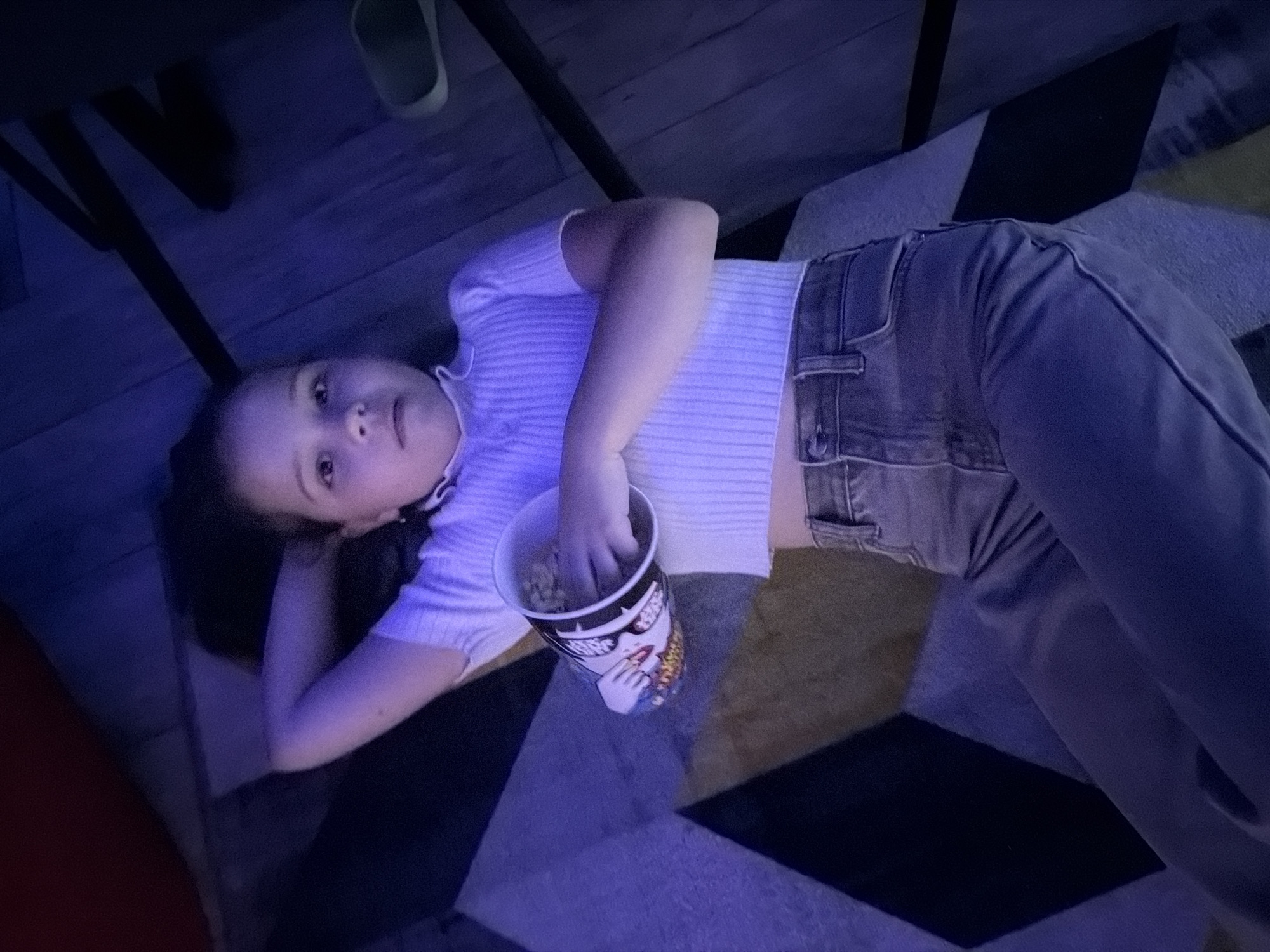 A young girl lies on her back on a patterned rug, her head resting on her arm. She is wearing a white, ribbed, cropped t-shirt and grey jeans. In her other hand, she holds a bucket of popcorn. The lighting in the room is predominantly blue, casting an ethereal glow on the scene. The room appears to be dimly lit, possibly indicating a movie-watching setting. The background consists of wooden panels and dark furniture, contributing to the cozy atmosphere. The image does not provide any specific details that would allow for identification of the location within Votkinsk, Russia.