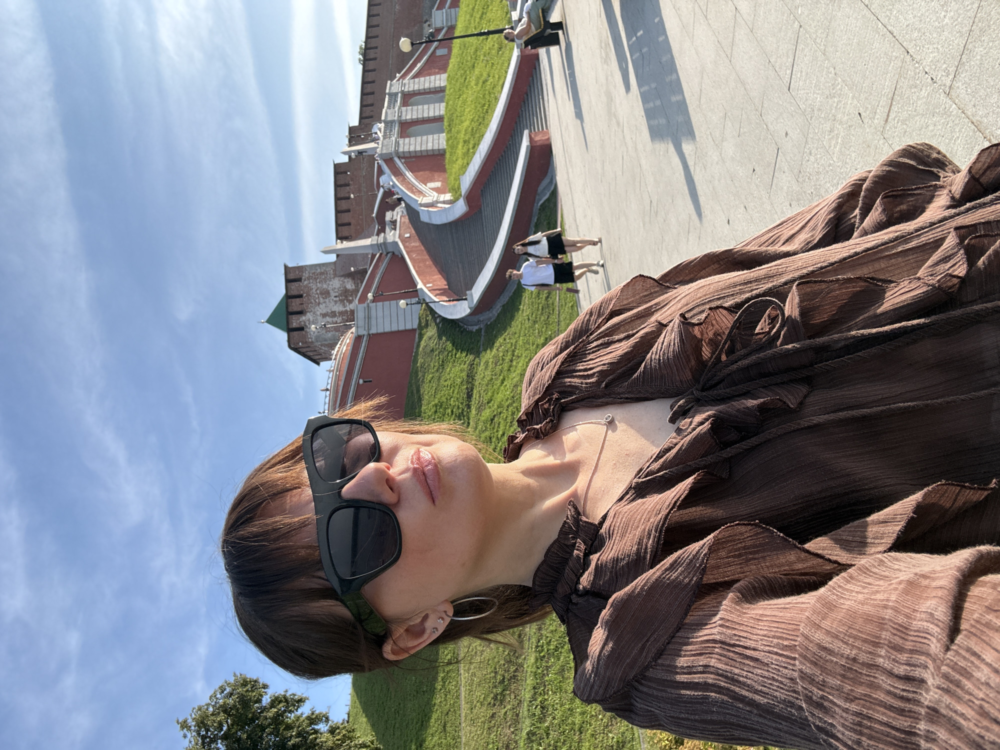 This media file shows a woman taking a selfie outdoors. She is wearing large, dark sunglasses and a brown, ruffled top. A thin silver necklace is visible around her neck. She is looking at the camera with her mouth slightly open. In the background, a large red brick structure with a green turret is visible, along with a wide, paved walkway and a grassy slope. Two other women in dark skirts and white tops are walking on the walkway. The scene is brightly lit, suggesting it is daytime. The sky is blue with some wispy clouds. The architecture and style of the clothing suggest a European location, possibly Russia as indicated by the location context. The overall mood appears casual and relaxed, as if enjoying a sunny day out. There is no visible text in the image.