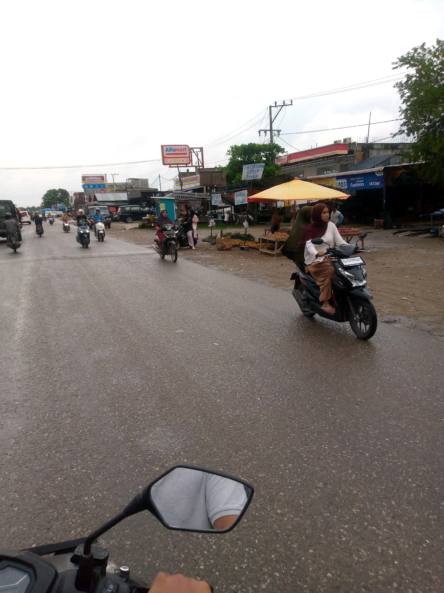 The image depicts a street scene in Gampongpaya, Indonesia, likely on a cloudy day given the overcast sky and the wet appearance of the asphalt road. Multiple motorbikes are present, with several individuals riding them and one parked. On the right side of the road, there is a woman wearing a hijab and a long-sleeved shirt with brown pants, riding a black motorbike. Adjacent to the road, a yellow umbrella shades a small stall, and further back, commercial buildings with signage, including "Alfamart" and "Indomaret," are visible. The presence of various shops and market stalls suggests this is a commercial or roadside market area. The overall impression is that of a busy, active street with daily commerce and transportation.