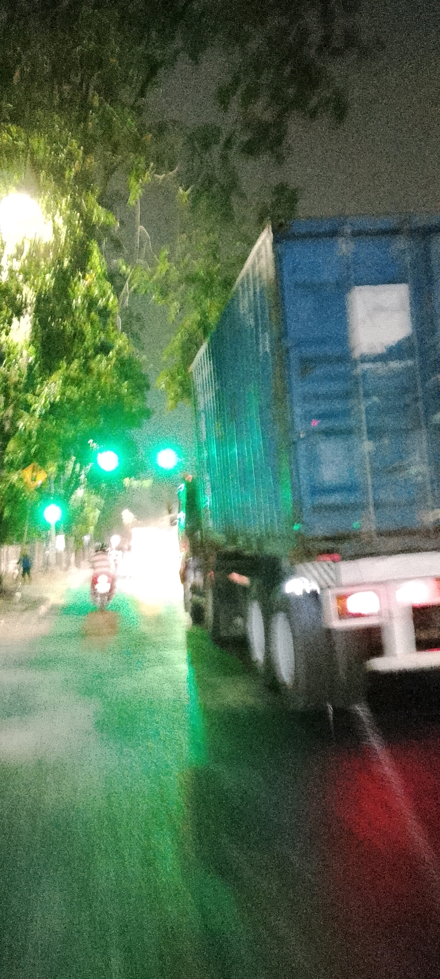 A semi-trailer truck with a blue shipping container is stopped at a traffic light in Bekasi, Indonesia, on a wet road at night. Green traffic lights are illuminated. A motorcycle is visible behind the truck, and other indistinct figures are present. Trees line the street, and a streetlight casts a bright glow on the foliage. The scene is characterized by motion blur and low light conditions, indicating a possible long exposure photograph or low-light camera setting.