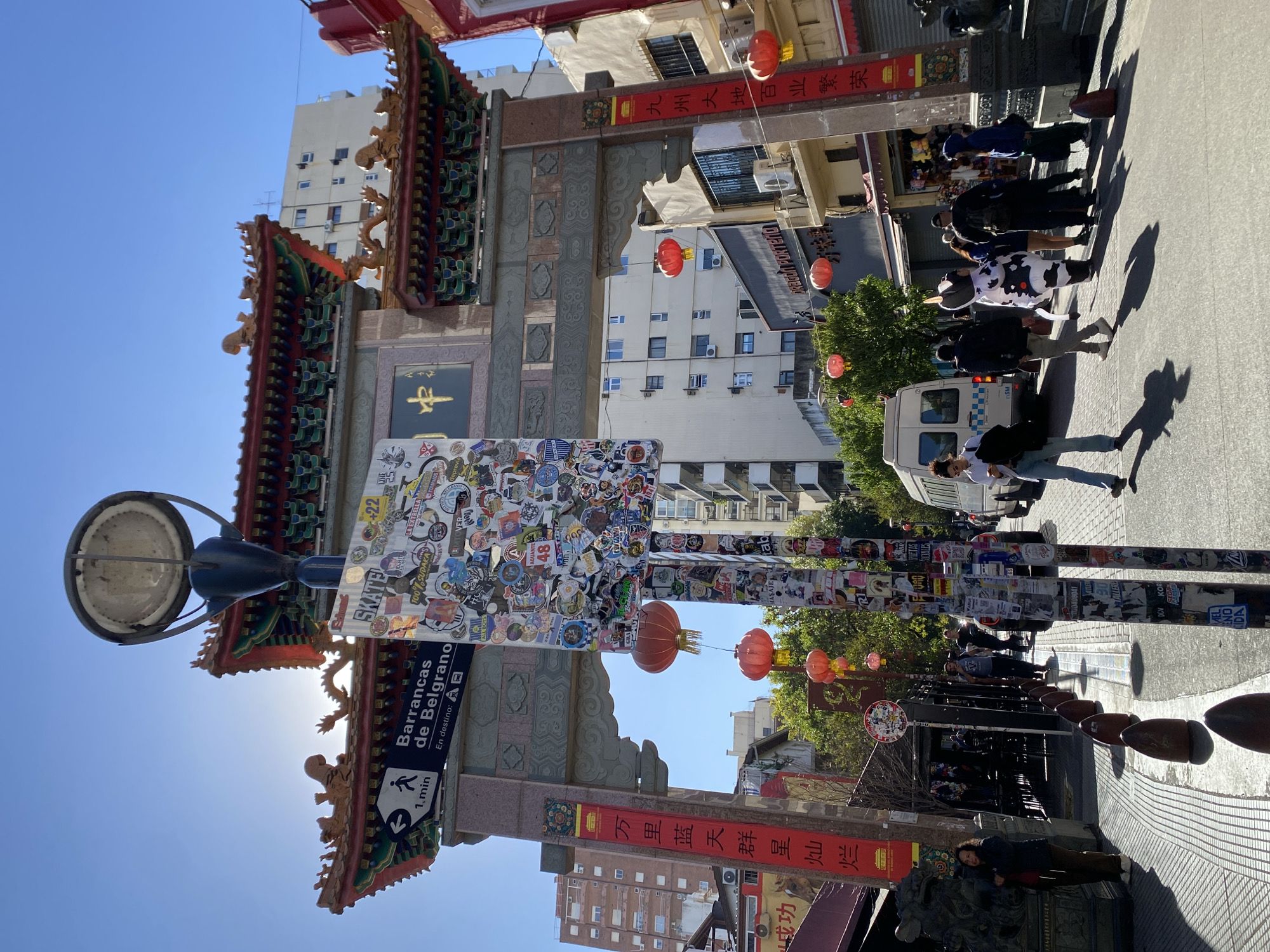 This image captures a vibrant street scene in the Chinatown district of Barrancas de Belgrano, Buenos Aires, Argentina, under a clear blue sky. A large, ornate Chinese archway (paifang) with intricate red and green tiled roofs and golden decorative figures dominates the foreground, signifying the cultural entrance to the area.
The street is lively with pedestrians walking in bright sunshine, evidenced by the long shadows cast on the paved ground, suggesting either mid-morning or late afternoon. Among the people, one person stands out in a distinctive cow-print costume. Red Chinese lanterns are strung decoratively across the street, enhancing the festive atmosphere.
A prominent street lamp post and an adjacent pillar are densely covered with a diverse collection of stickers, displaying urban art, various logos, and text, including "SKATE", "ALL BOYS", and "100% SUDAKAS". A directional sign on the lamp post reads "Barrancas de Belgrano En destino: 1 min", confirming the location and indicating a short walk to the destination. Multi-story urban buildings line the street, some with visible greenery from trees.
Banners hanging from the archway feature Chinese characters that translate to phrases like "The vast land of China, all industries prosper" and "Ten thousand li of blue sky, shining stars". A shop sign nearby reads "DRAGOA PORTENO". The scene is a dynamic blend of traditional cultural elements and modern urban life.