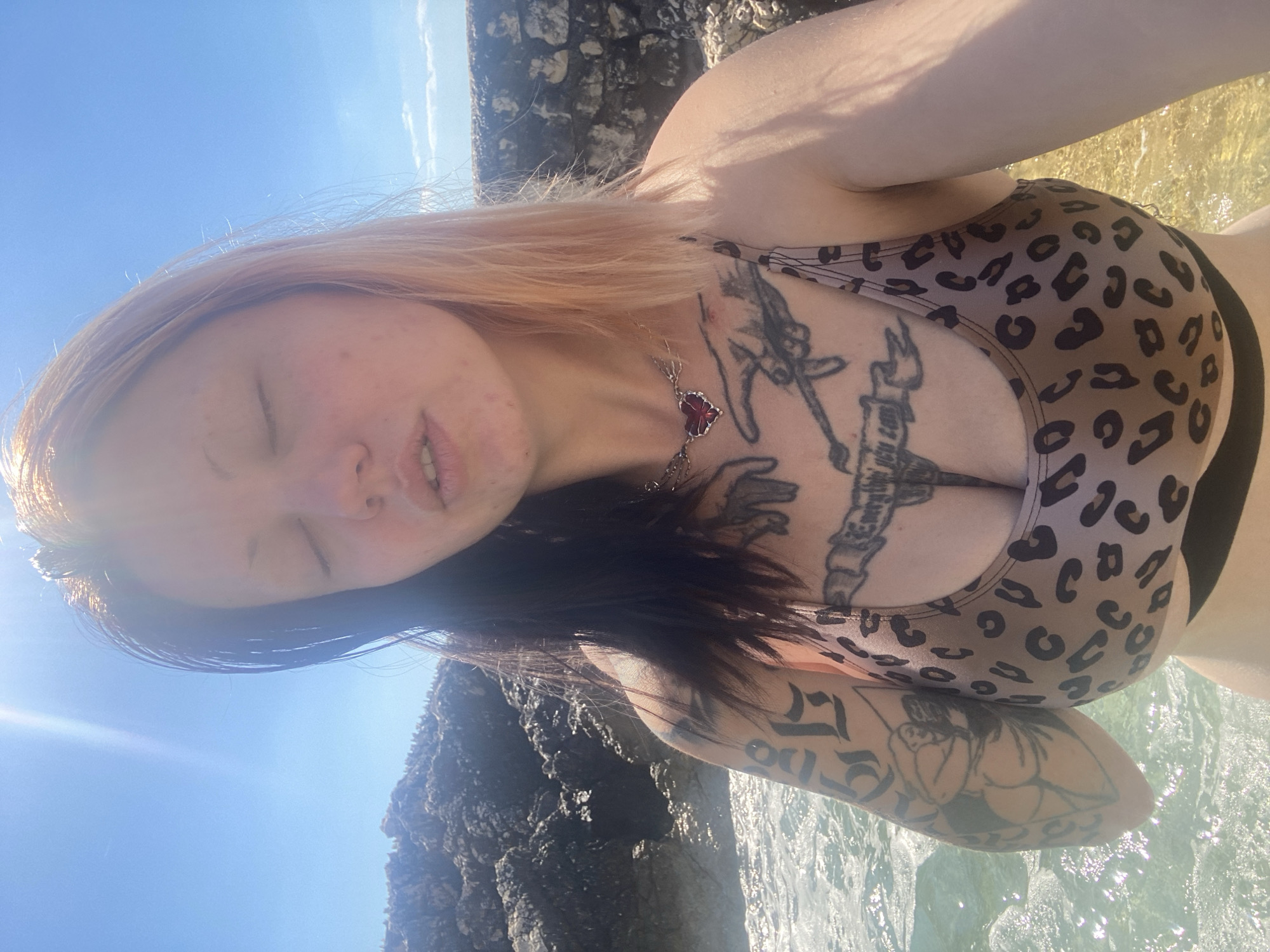 A light-skinned woman with a chest tattoo wears a cheetah-print bikini top. She has reddish-blonde hair on top and dark hair underneath. She wears a red necklace. She is standing in shallow, clear water near a rocky outcropping under a bright blue sky and sunlight, possibly in the coastal city of Bar, Montenegro.