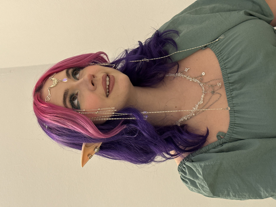 A woman with pink and purple hair poses, wearing pointed elf ears with silver hoops. Her makeup includes defined eyebrows and eyeliner. A decorative silver headband with a light purple jewel adorns her forehead. Strands of silver chains with crystals and small beads hang around her face and neck, styled as a necklace. She wears a green blouse with a gathered neckline and puffed sleeves, and the background is a plain white wall.