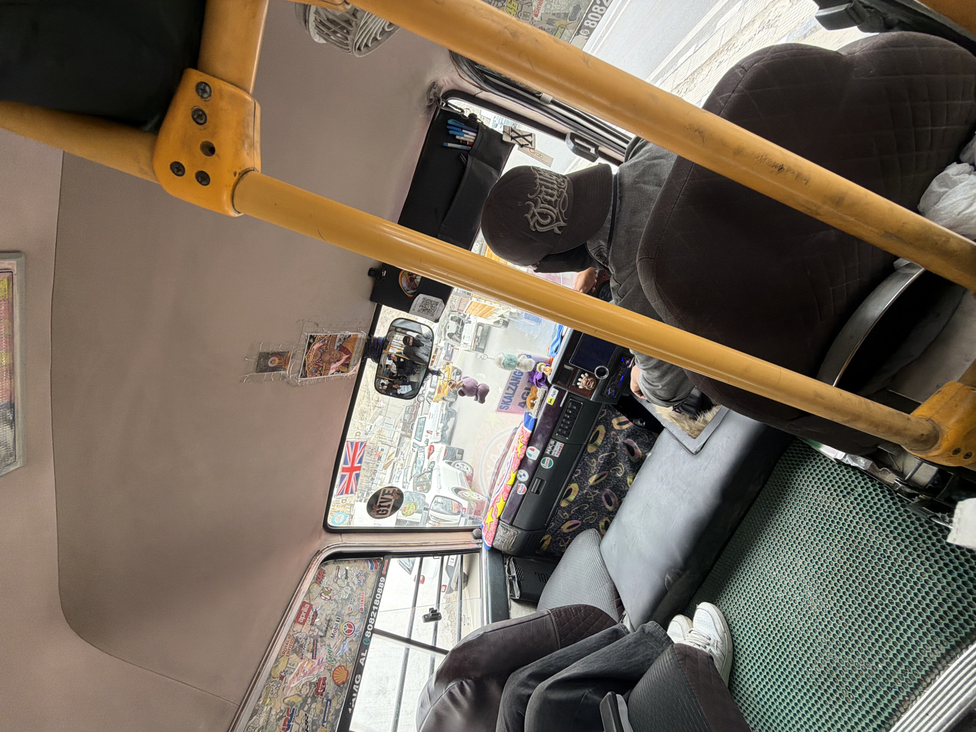 The image shows the interior of a bus, likely in Leh, India, as indicated by decorative elements common in local transportation. The scene is viewed from the perspective of a passenger, looking towards the driver and the front of the bus.
In the foreground, yellow metal safety bars frame the view. To the left, a passenger's legs are visible, wearing dark pants and white sneakers, with their feet resting on a green textured mat. A dark brown seat is partially visible.
The driver, wearing a grey jacket and a dark baseball cap with white lettering, is seated at the wheel. Their face is partially obscured by the cap and the angle of the shot. The dashboard is heavily decorated with stickers and various items. A prominent sticker reads "SKALZANG". A tablet-like screen is visible on the dashboard.
The interior of the bus is adorned with numerous stickers on the ceiling and window frames, featuring logos like Shell, Mobil, Yamaha, and a Union Jack flag. There are also small religious images, possibly Buddhist thangkas, and other decorative trinkets hanging from the ceiling and dashboard.
Through the windshield, a street scene is visible, showing other vehicles, including cars and what appear to be local taxis or buses, suggesting a busy urban or semi-urban environment. The lighting indicates it is daytime, possibly overcast. The overall impression is of a personalized and well-decorated interior of a public transport vehicle.