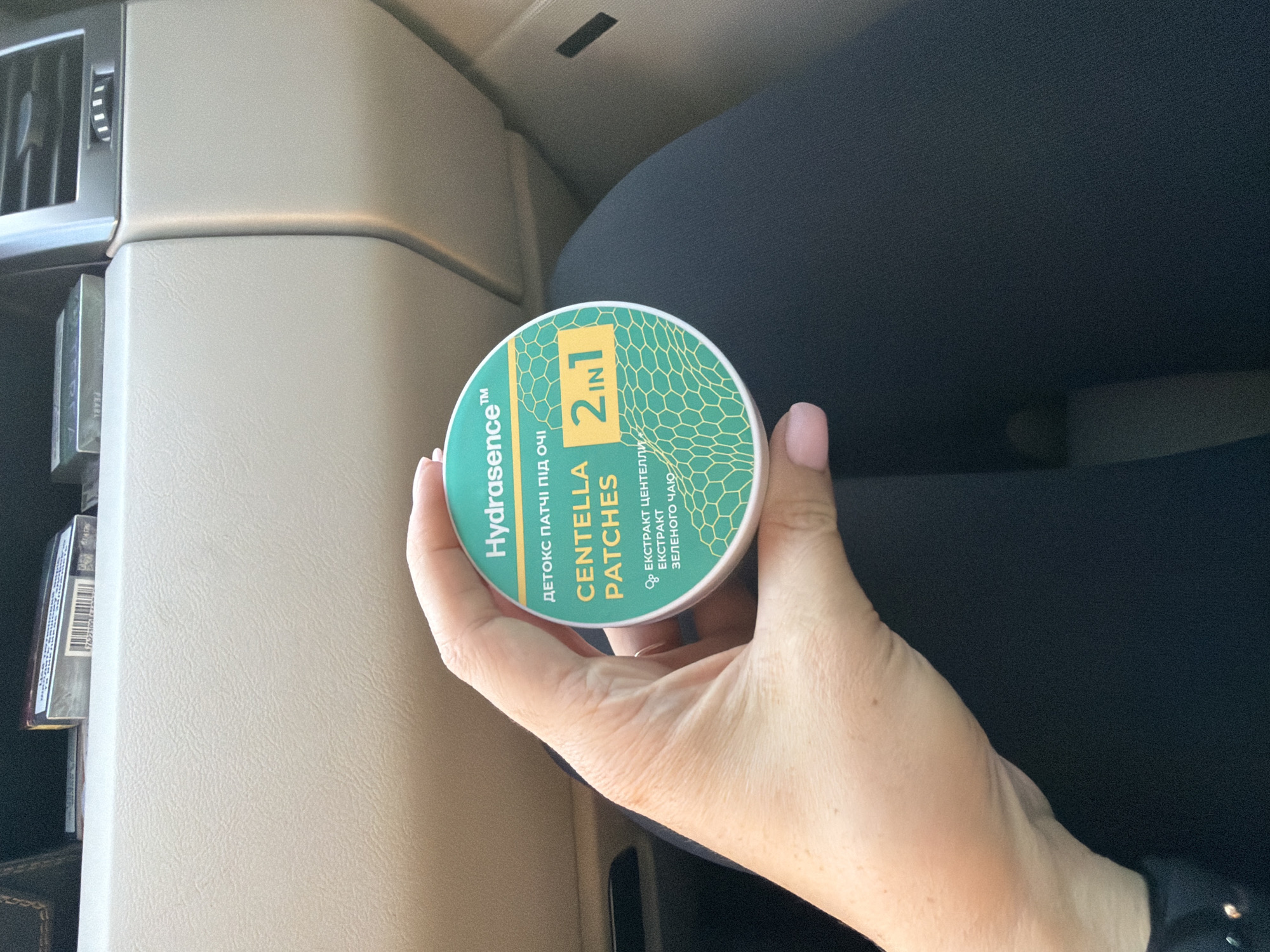 A hand holds a round, green and white container of "Hydrasense Centella Patches 2 in 1" in the foreground. The container has text in Ukrainian and English, as well as a honeycomb-like pattern. In the background, the interior of a car is visible, including a beige dashboard, a black air vent, and a partially obscured pack of cigarettes in a compartment. The person's hand is resting on dark fabric, likely clothing. The overall setting appears to be inside a vehicle.
